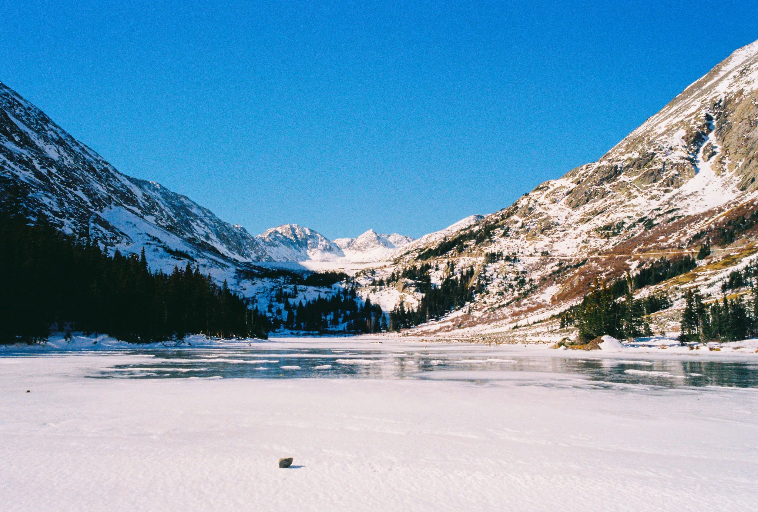 Snow-covered landscape of a mountain valley with a frozen lake, surrounded by tall snow-capped mountains and pine trees under a clear blue sky.