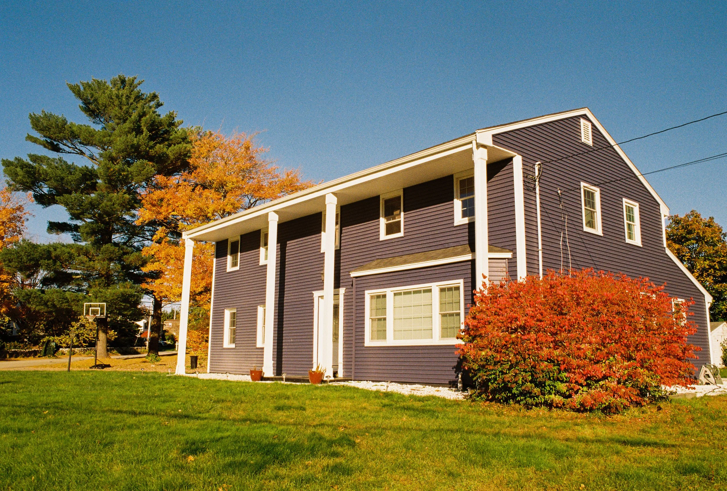 A two-story house with purple siding and white trim, surrounded by trees with autumn leaves, on a sunny day.