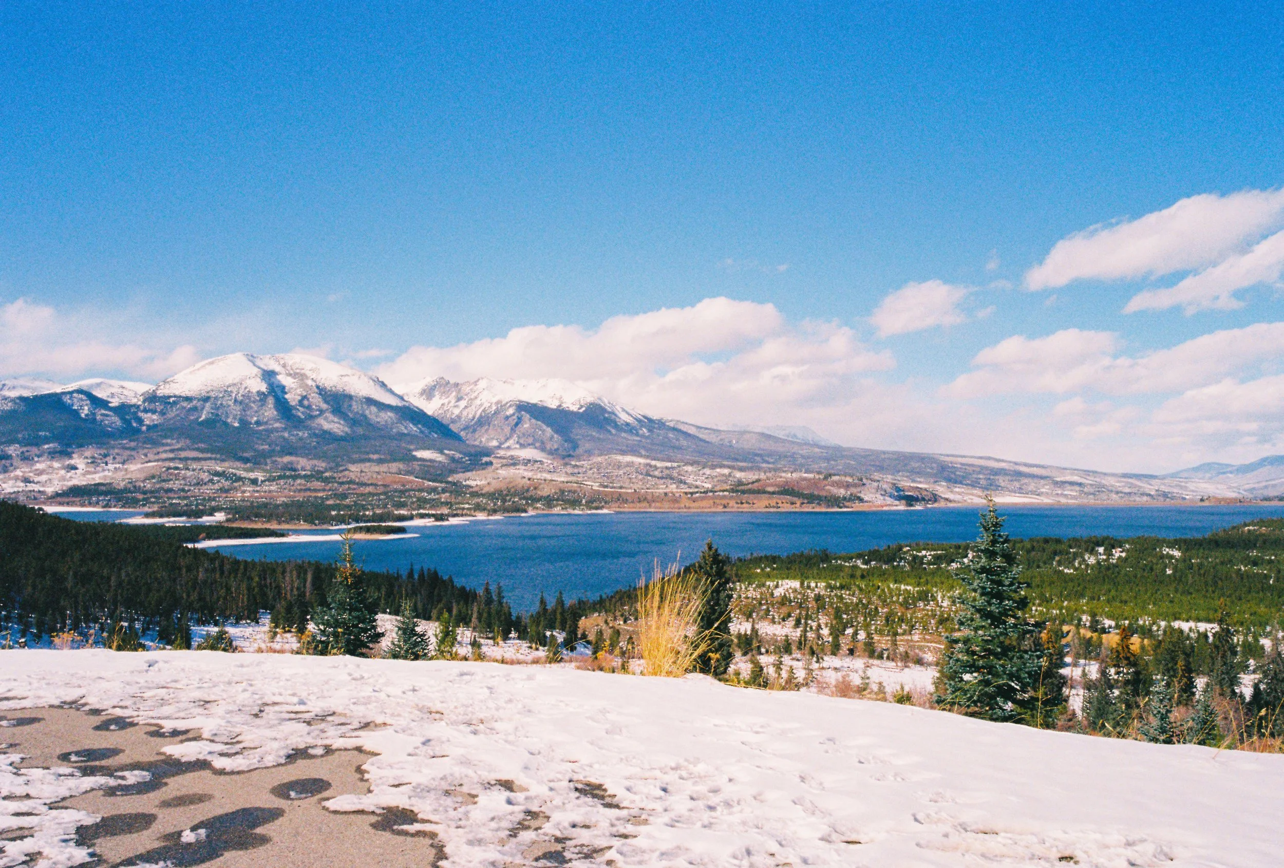 Snow-covered ground with a large lake, green forests, snow-capped mountains, and a bright blue sky with some clouds.