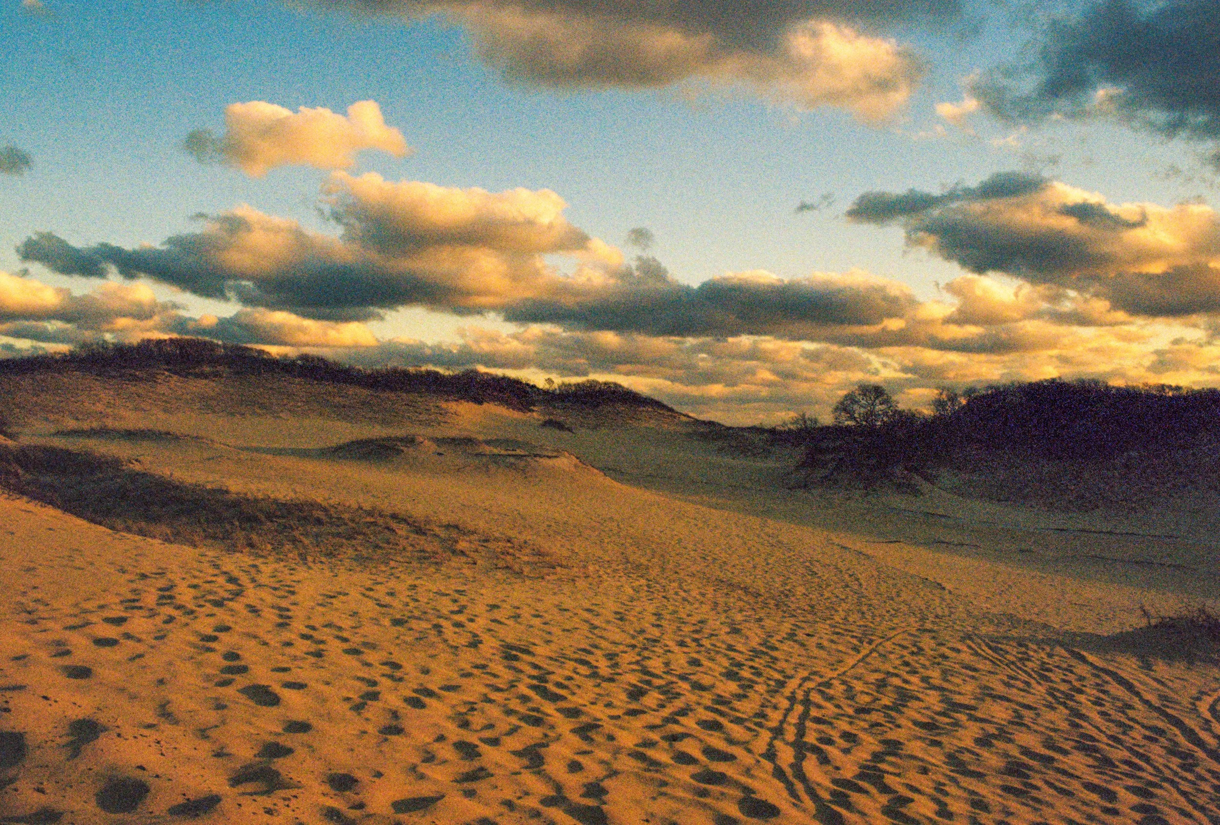 Sand dunes with footprints and tire tracks under a partly cloudy sky at sunset.