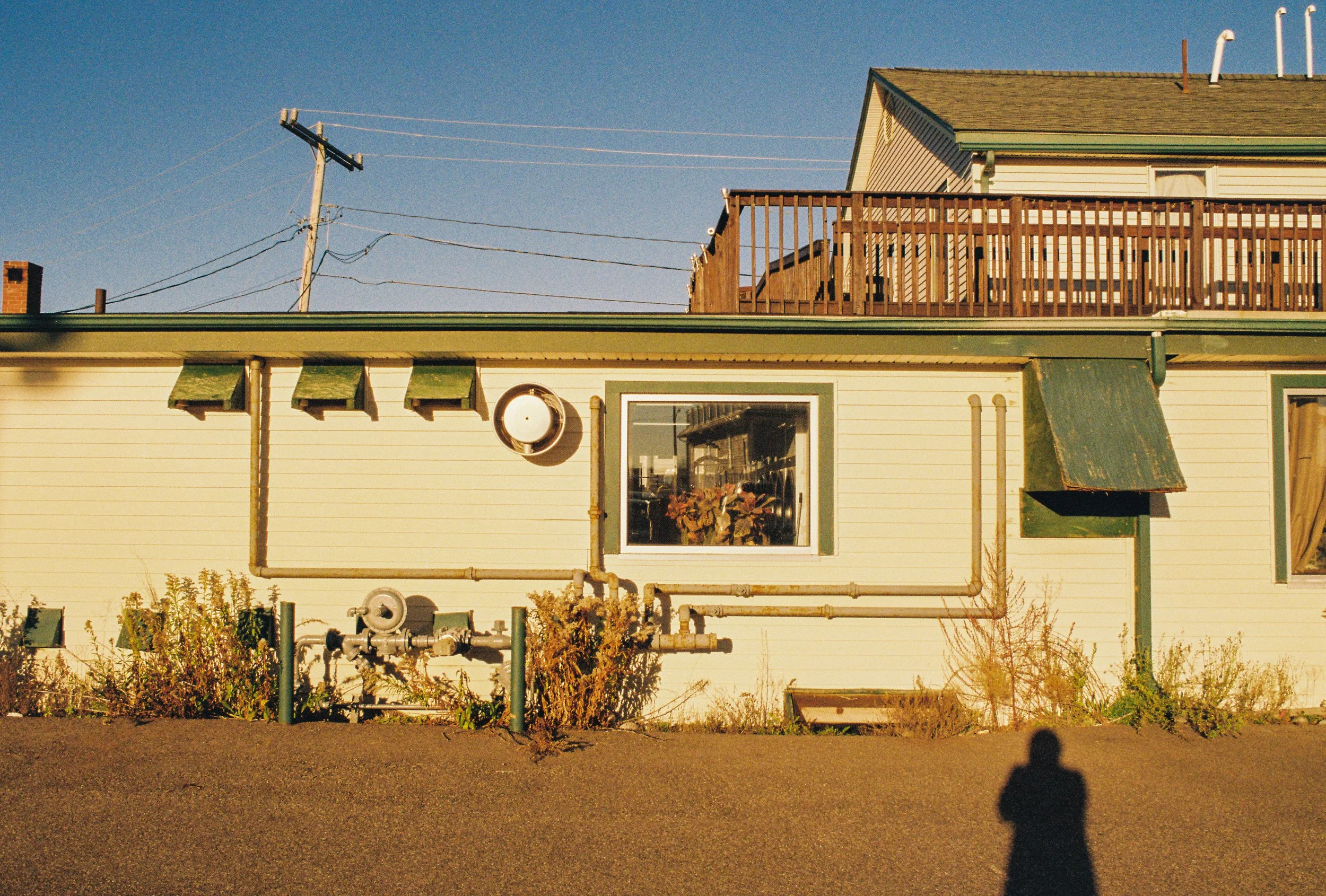 A beige house with green accents, a window with a plant inside, a wooden deck on the roof, and a shadow of myself taking the photo in the foreground, under a clear blue sky.