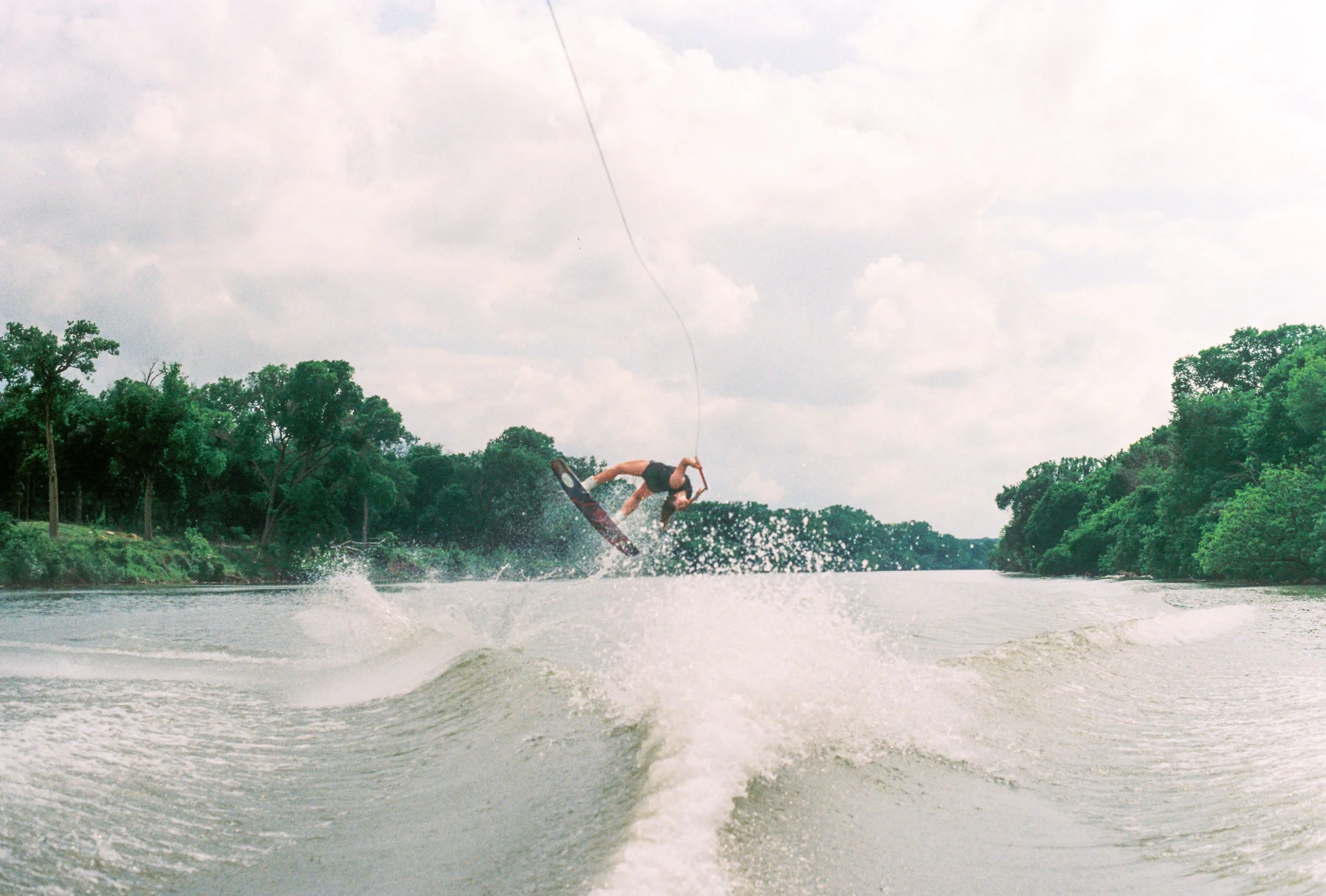 A woman performing a jump on a wakeboard on a river, with water spray and trees along the riverbank under a cloudy sky.