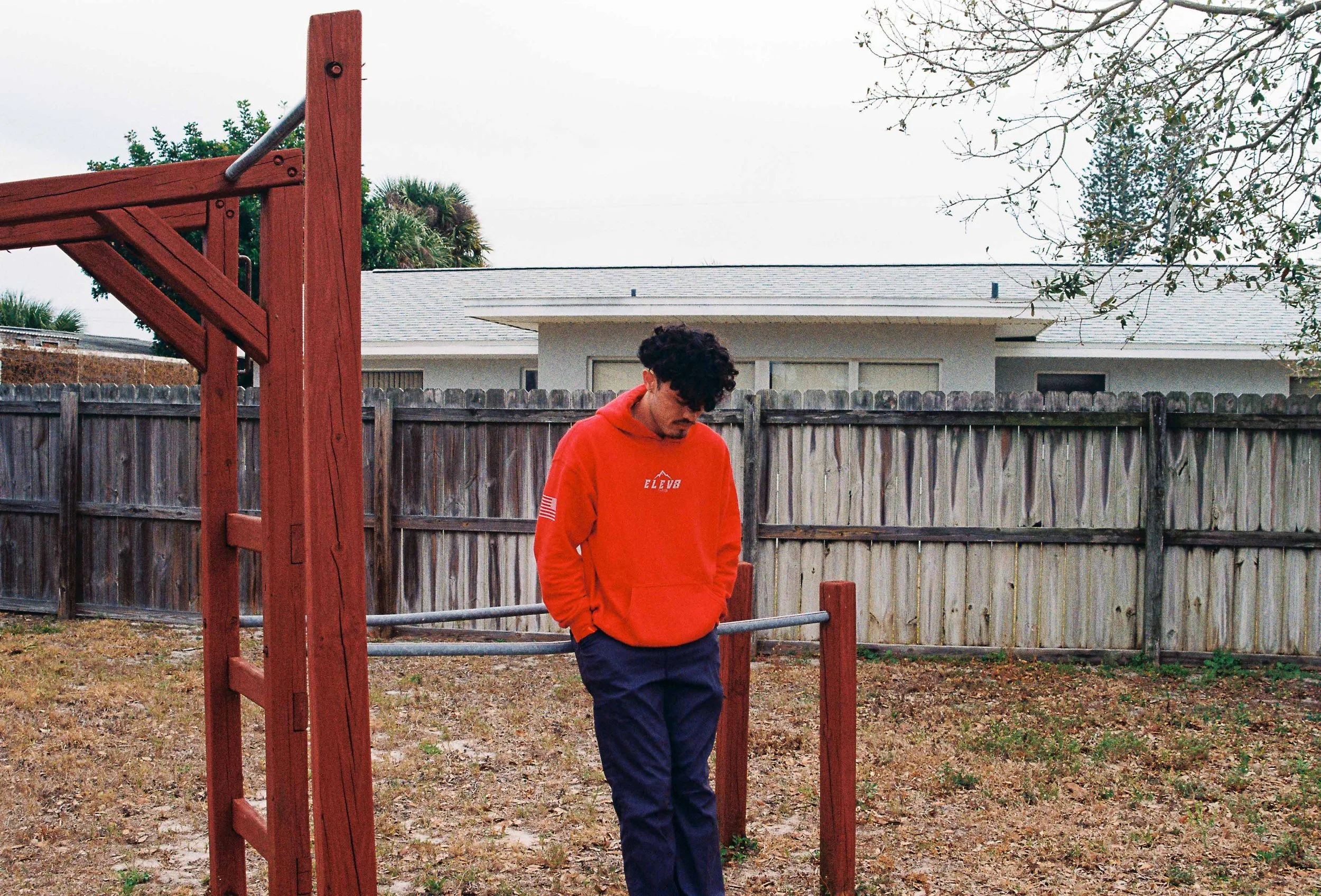A young man with curly hair and a mustache wearing a red hoodie and dark pants standing outdoors in a backyard, near a red wooden playground structure, with a wooden fence and a house in the background under an overcast sky.