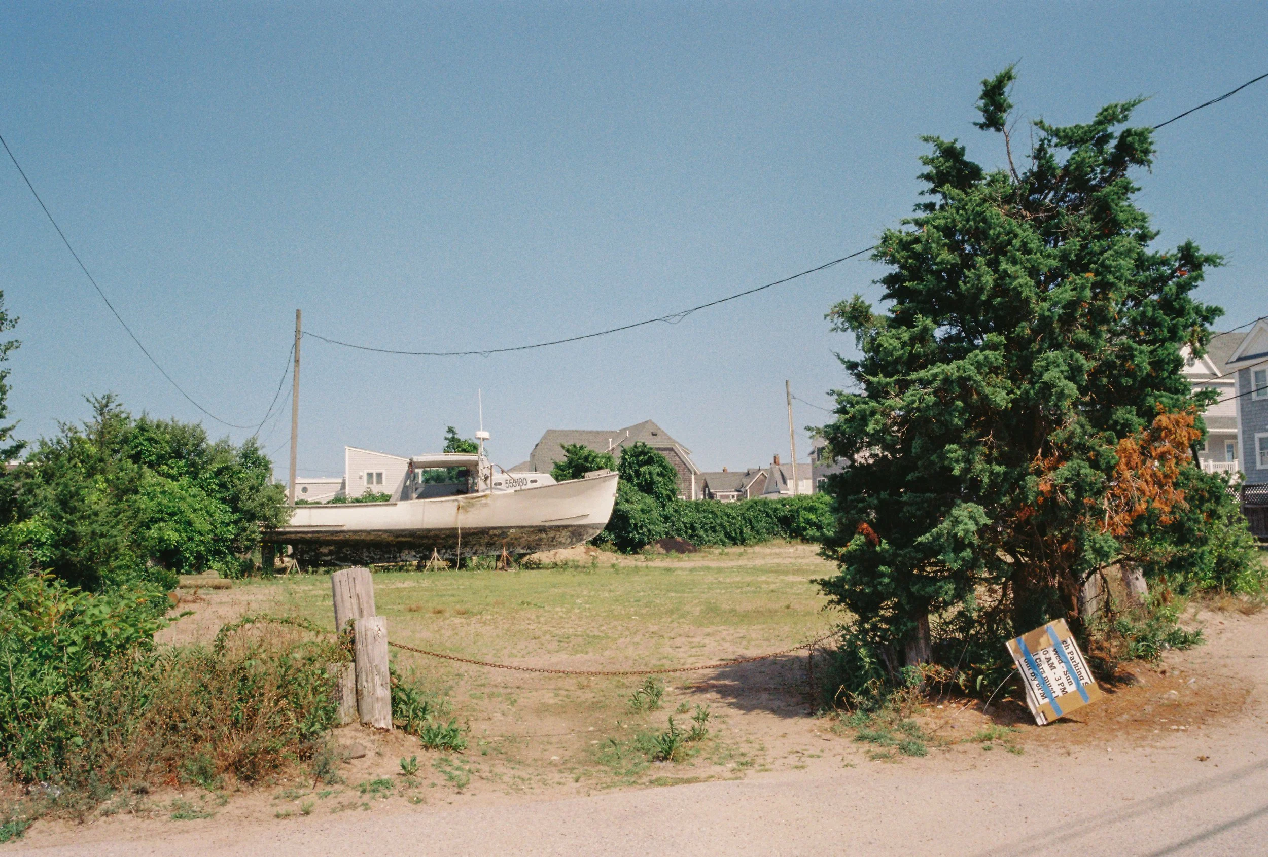 A boat on land with grass, bushes, a tree, and a sign near the tree, with houses in the background and power lines overhead.