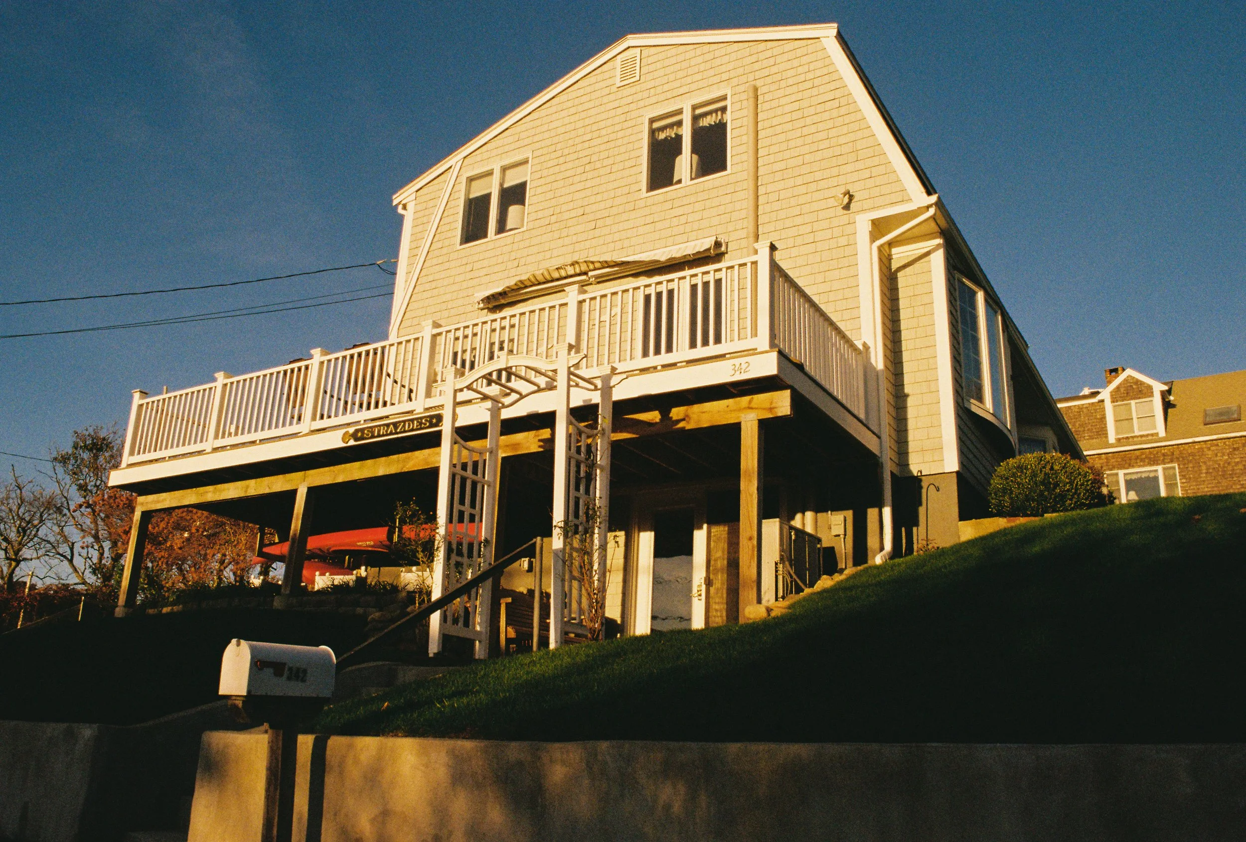 A two-story house elevated on stilts with a large white deck and railing, a set of stairs leading up to the deck, a small boat underneath, and a mailbox in the foreground, all set during sunset.