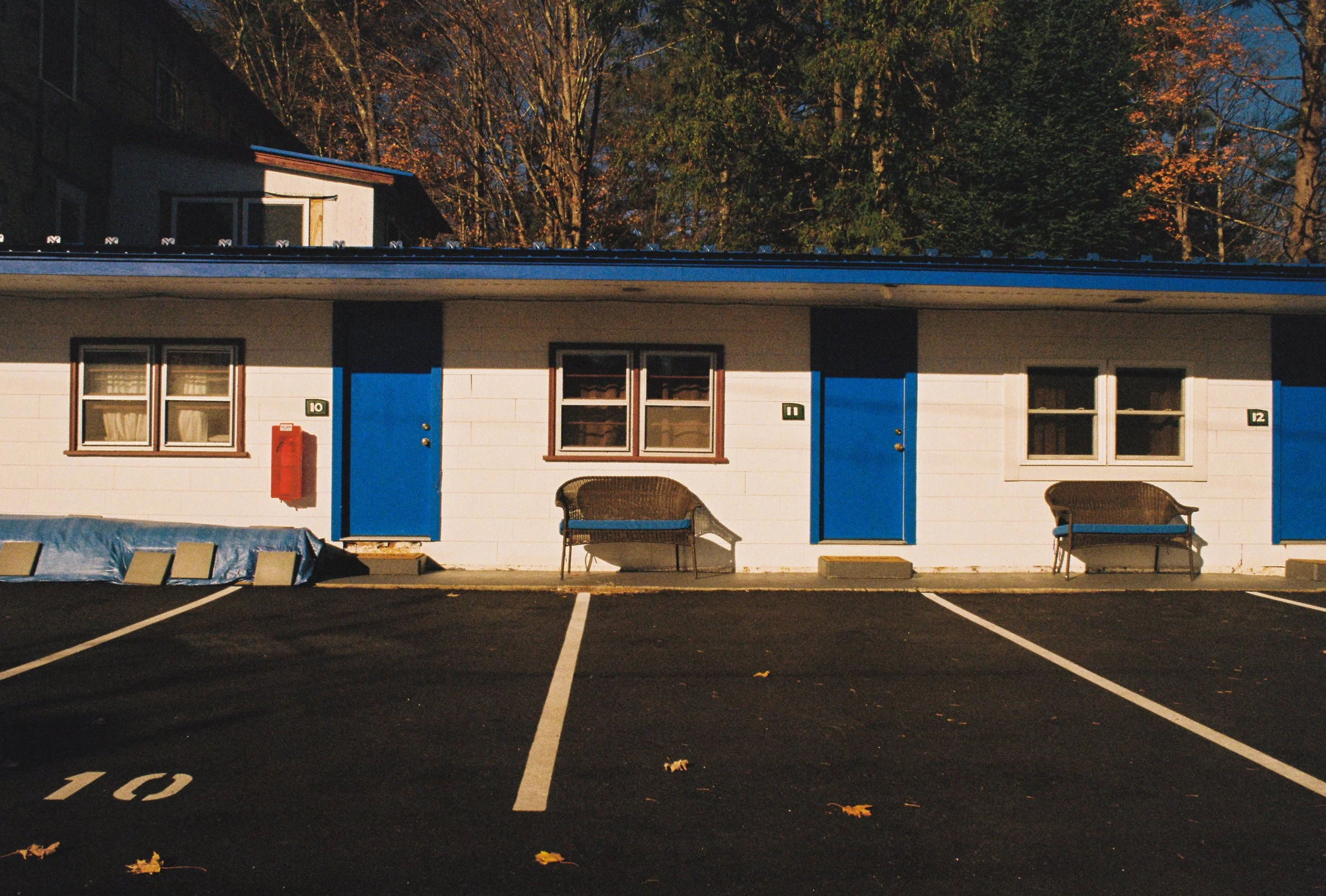 A motel with parking spaces in front, two benches outside, and a red fire alarm box on the wall. The building has white siding with blue doors and window shutters, and trees with fall leaves are in the background.