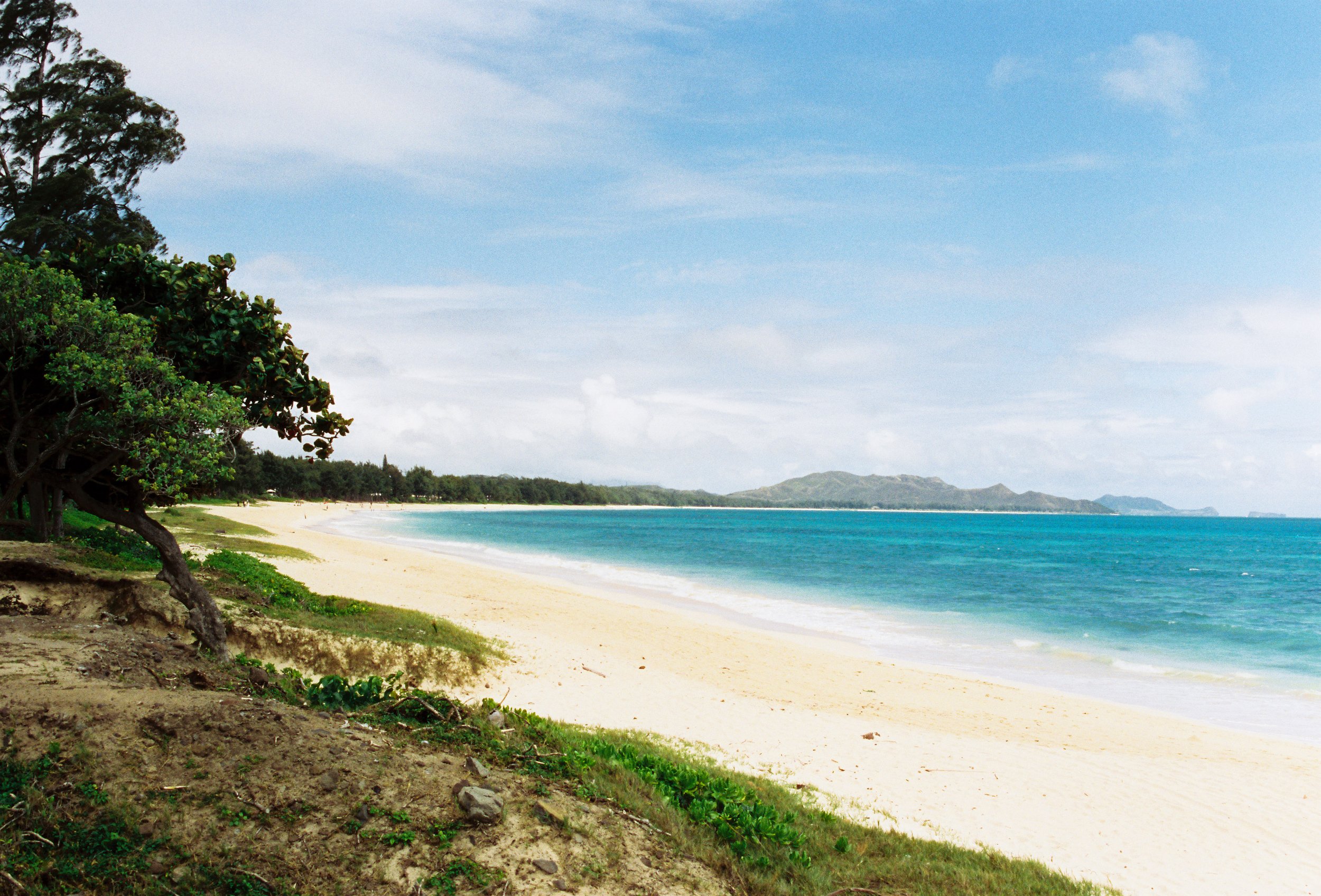 Tropical beach with sandy shore, turquoise water, green trees, and distant mountains under a partly cloudy sky.