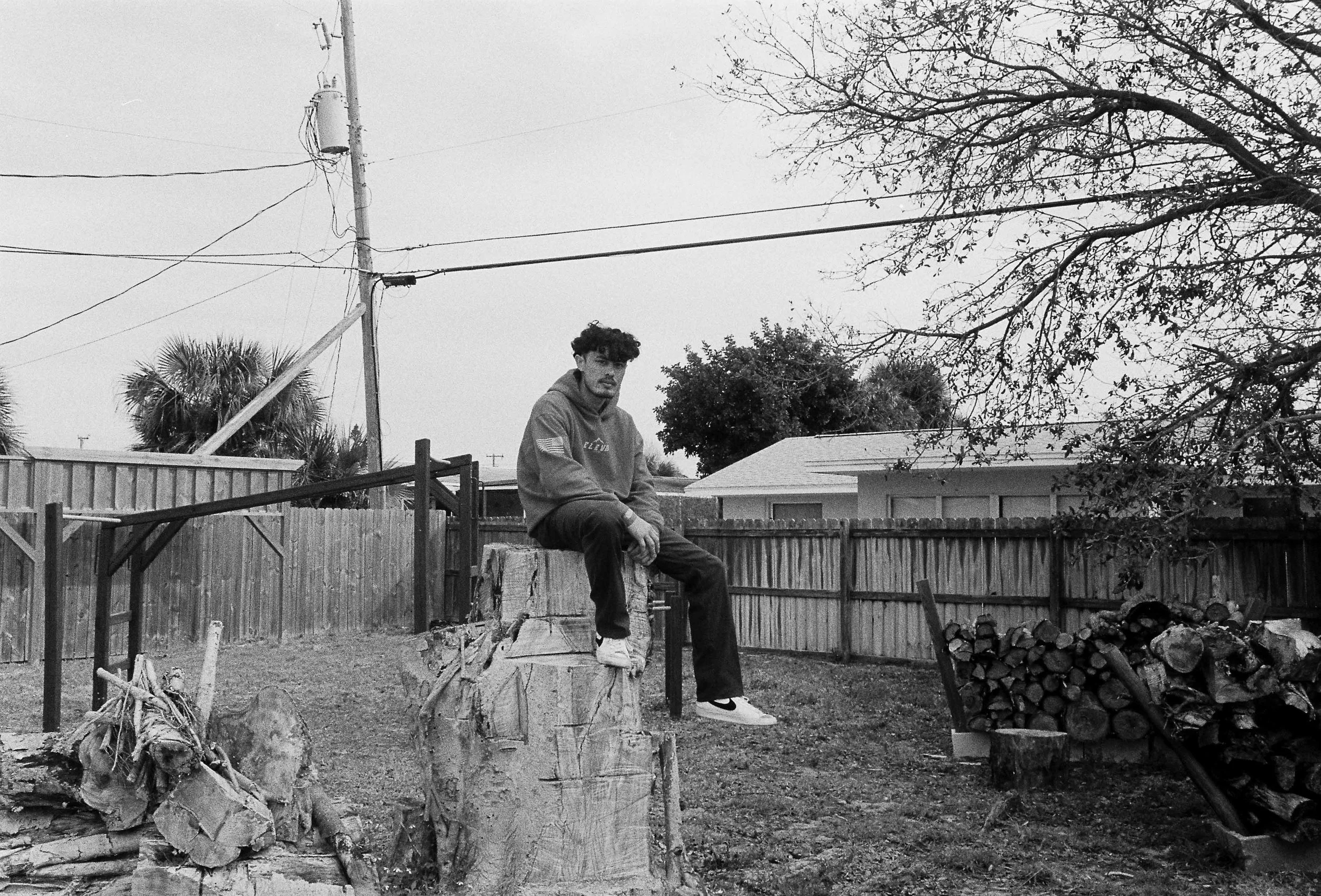 A young man with curly hair sitting on a wooden bench in a backyard, surrounded by trees, wooden fence, and stacked logs, in black and white.
