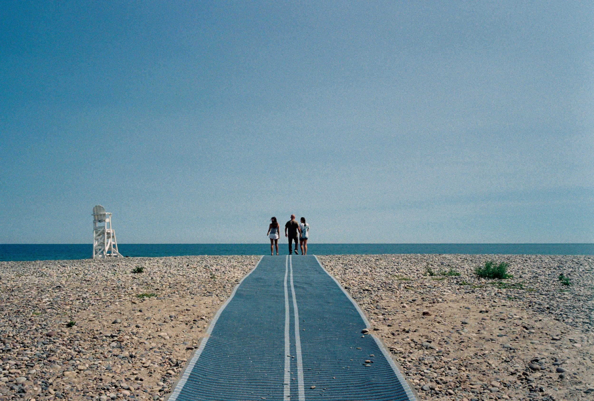 Three people walking along a pathway on a beach towards the ocean, with a lifeguard tower on the left and a clear blue sky above.