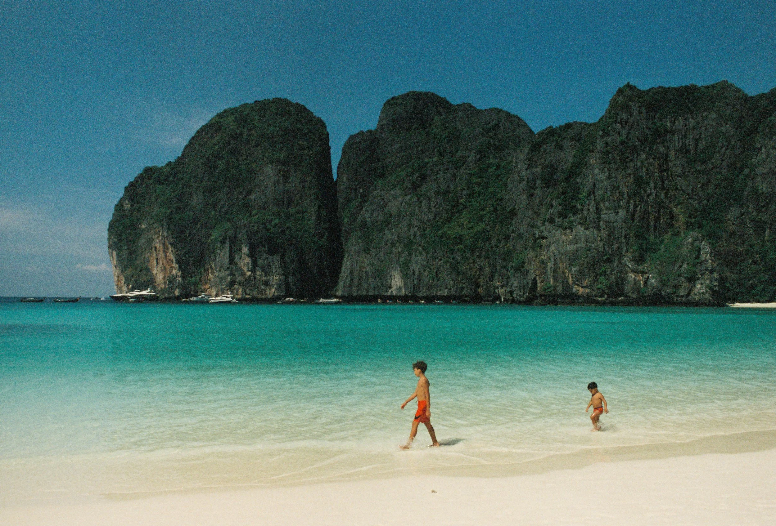 Two children playing in the shallow water on a tropical beach with large rocky cliffs in the background.