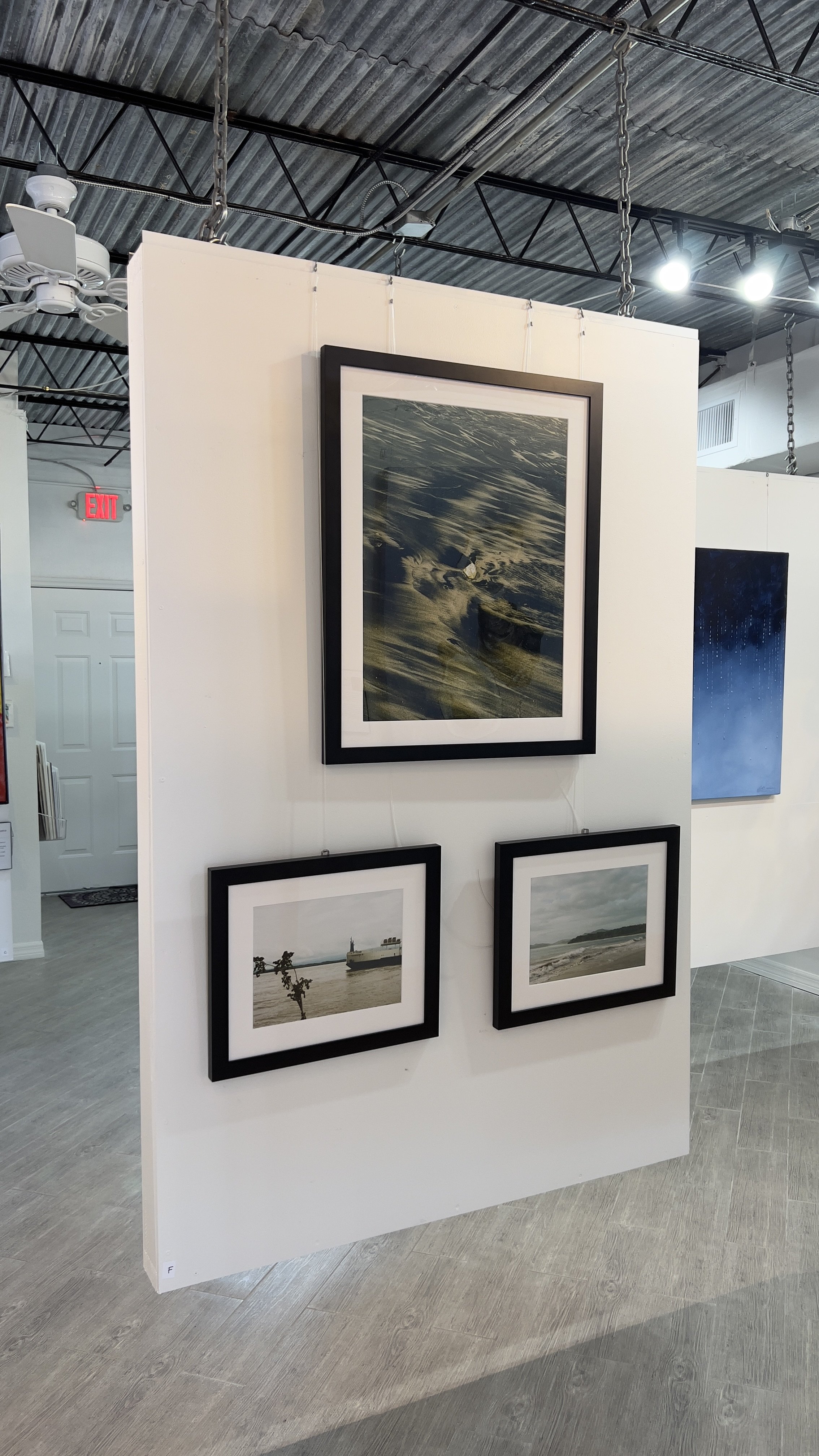 Three framed photographs of coastal scenes displayed on a white partition in an art gallery.