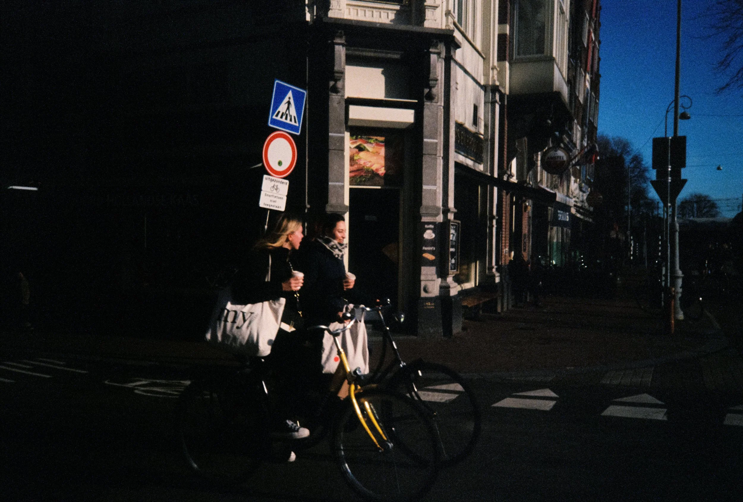 Two women walking next to each other, one riding a bicycle, at night in an urban area with buildings and street signs visible.