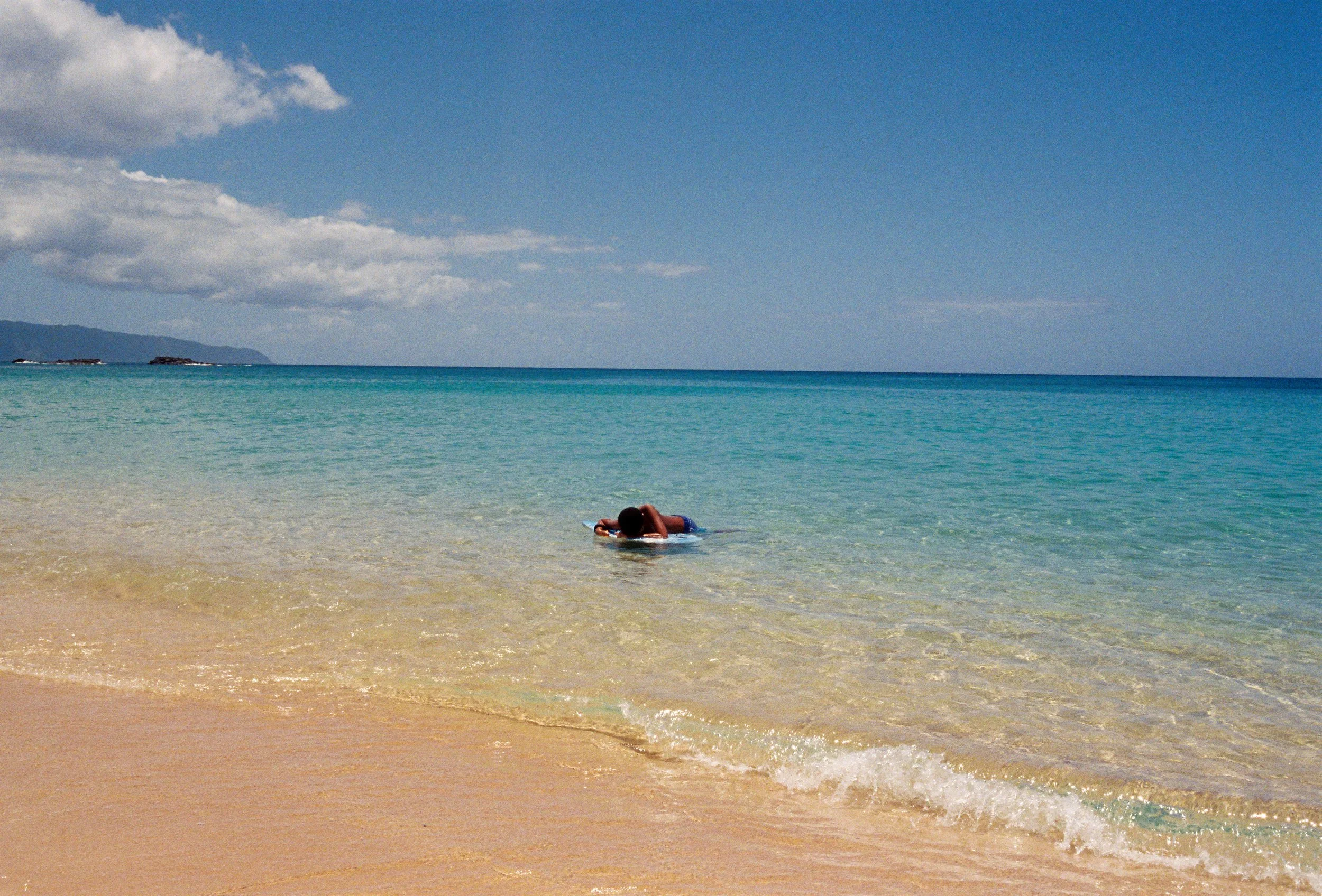 A person lying on a surfboard in shallow, clear ocean water near a sandy beach, with a distant coastline and partly cloudy sky in the background.