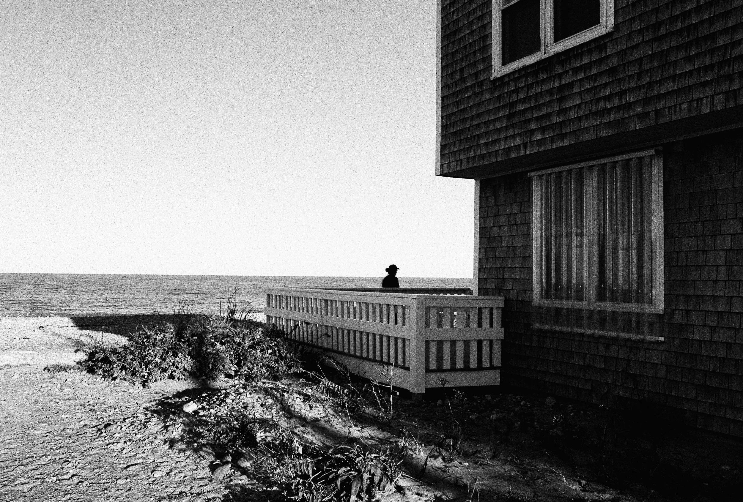 A person standing on a balcony overlooking the ocean, with a house on the right and a bush in the foreground, in black and white.