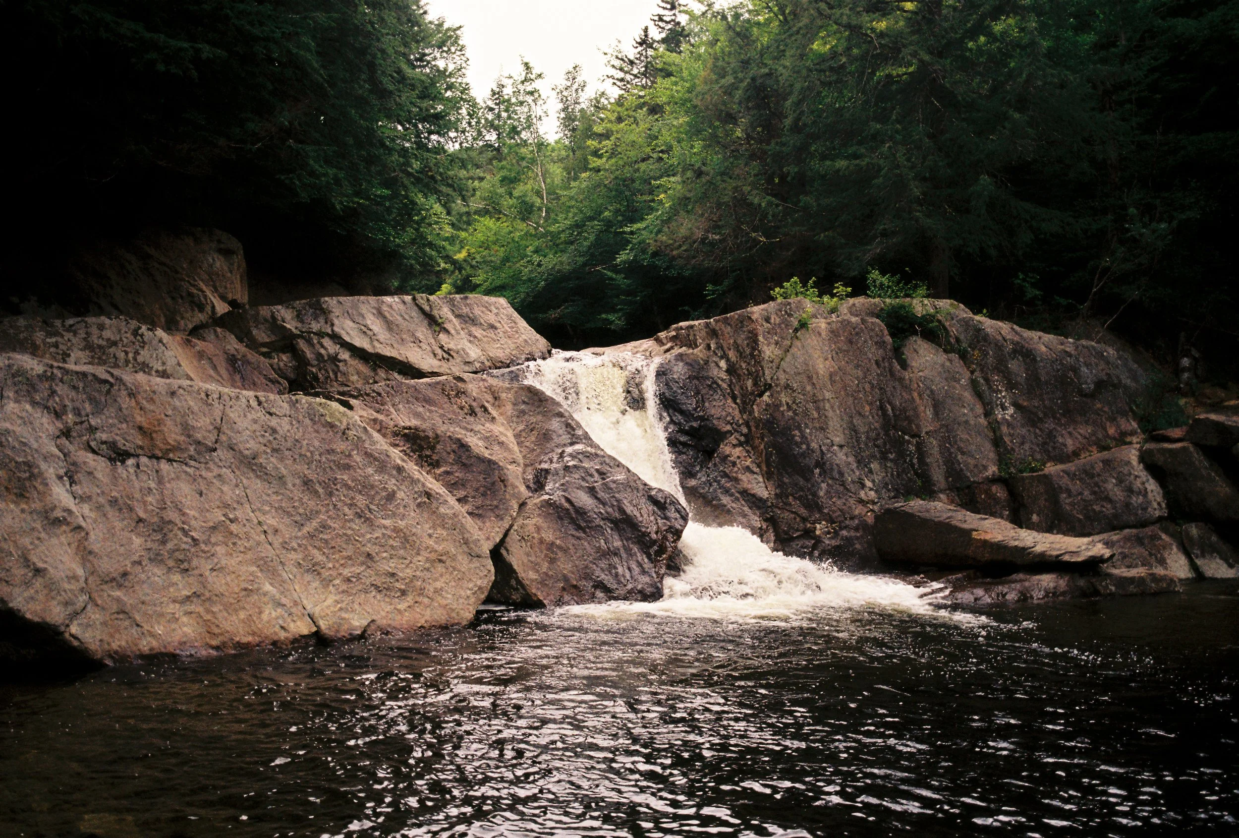 A small waterfall flowing over large rocks into a dark river surrounded by green trees.