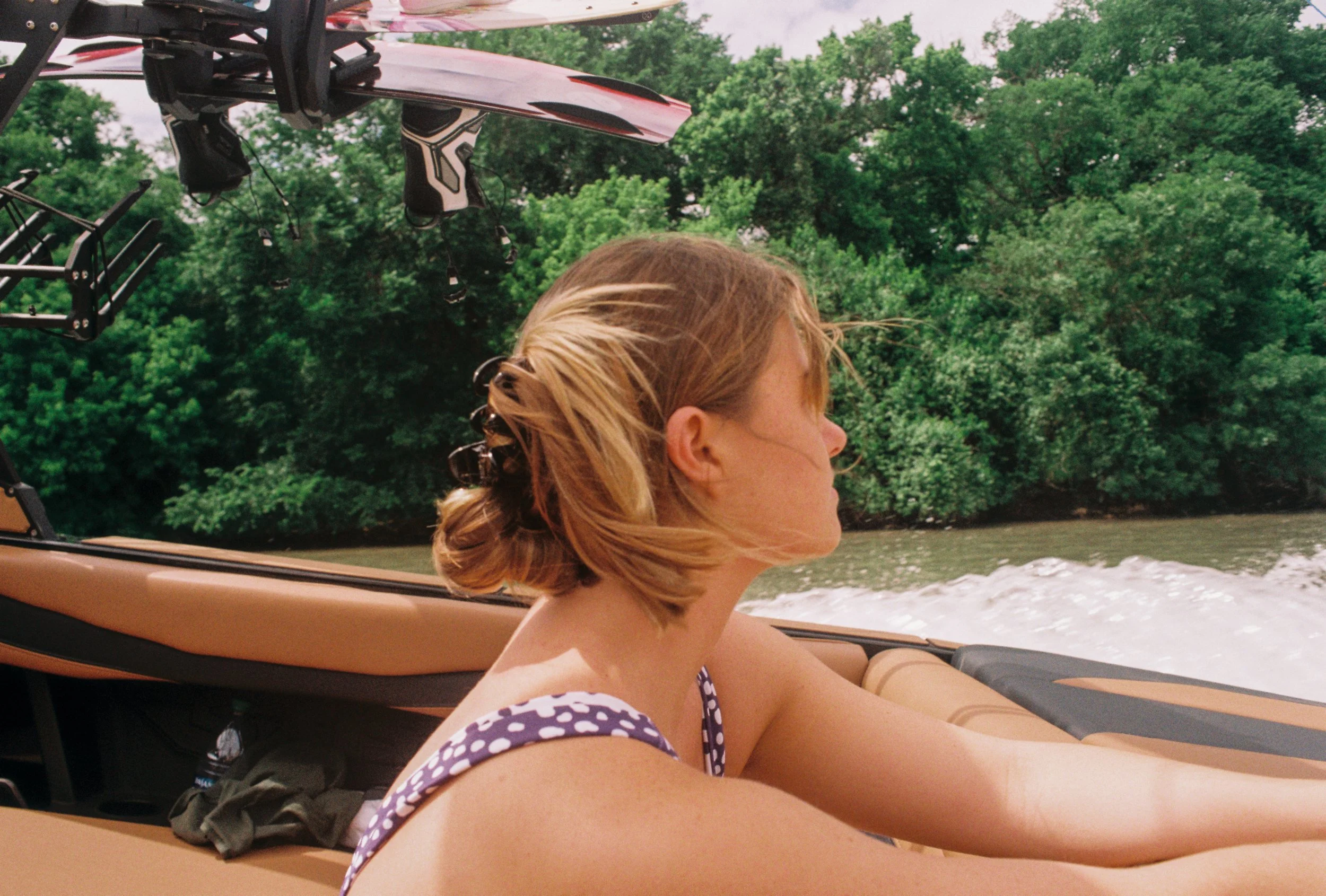 A woman with blonde hair in a ponytail wearing a polka dot swimsuit, sitting in a boat on the water, facing sideways with trees in the background.