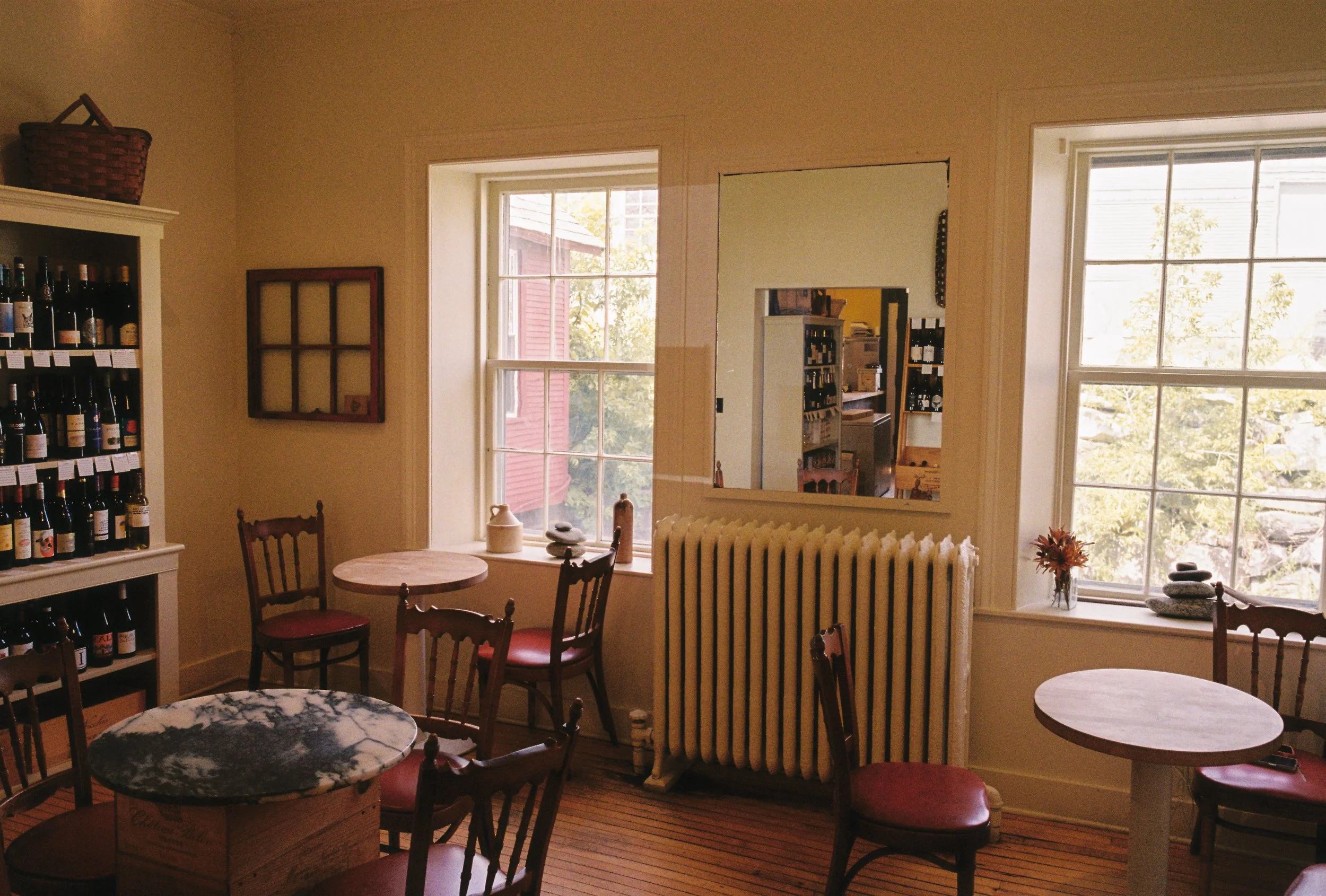 Interior of a cozy cafe or wine shop with white walls, wooden flooring, two large windows, a radiator, a mirror on the wall, a shelf of wine bottles, and small round tables with red cushioned chairs.