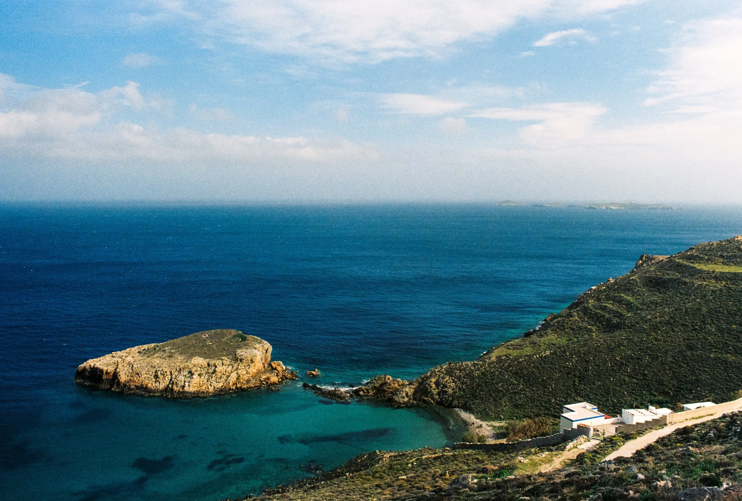 A coastal landscape with a small white building near a winding road, green hills, a rocky island, and the ocean extending to the horizon under a partly cloudy sky.