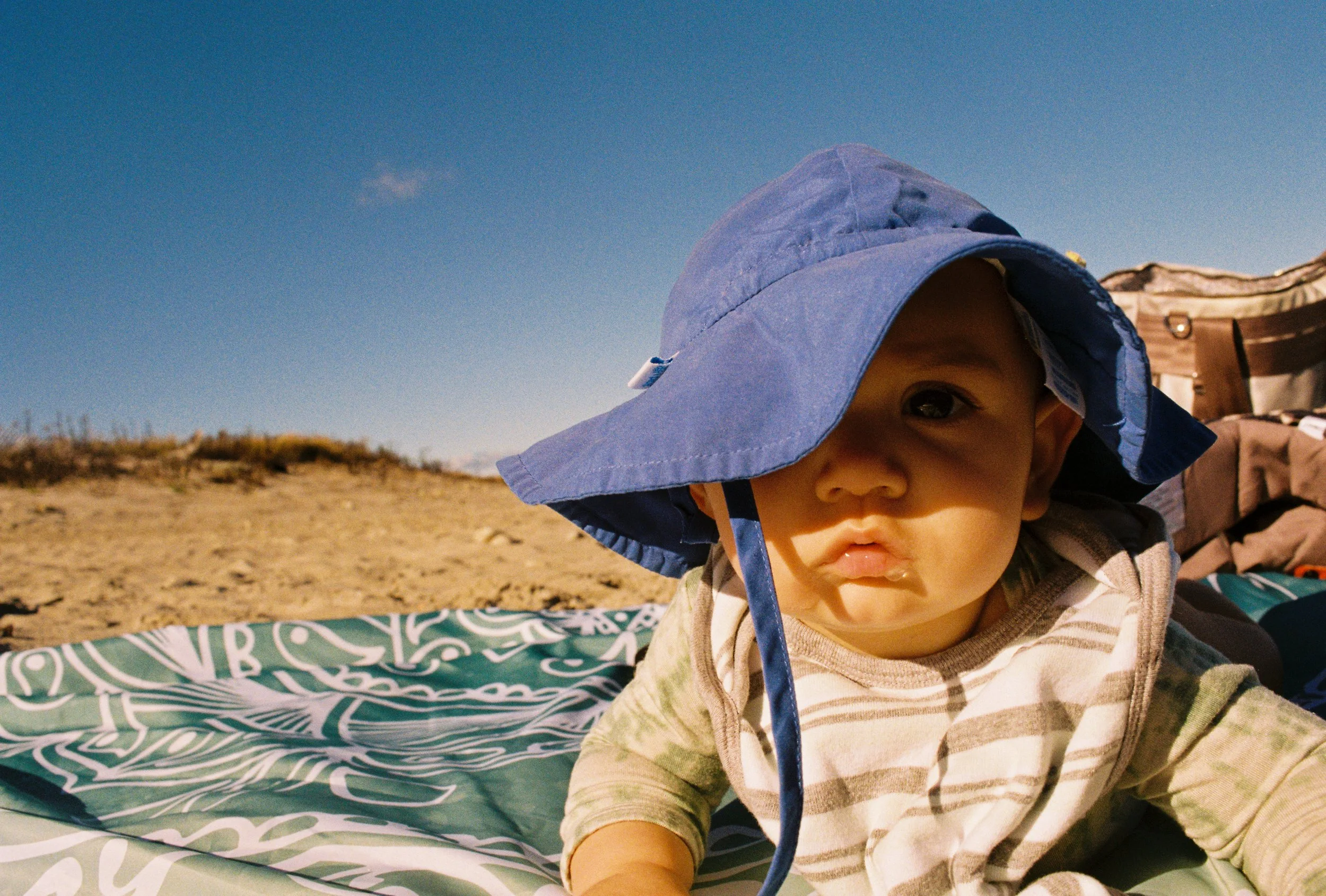 A young child wearing a blue sun hat and a striped shirt outdoors on a sunny day, lying on a patterned blanket on sandy ground.