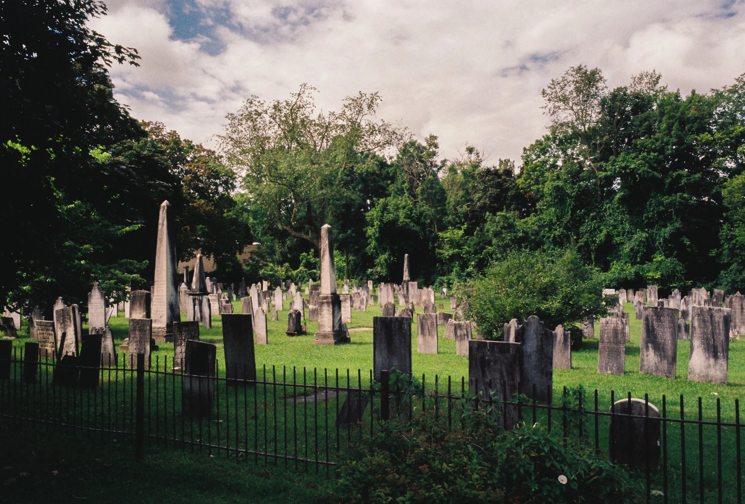 A cemetery with old, weathered tombstones surrounded by a black metal fence, green grass, and trees in the background under a partly cloudy sky.