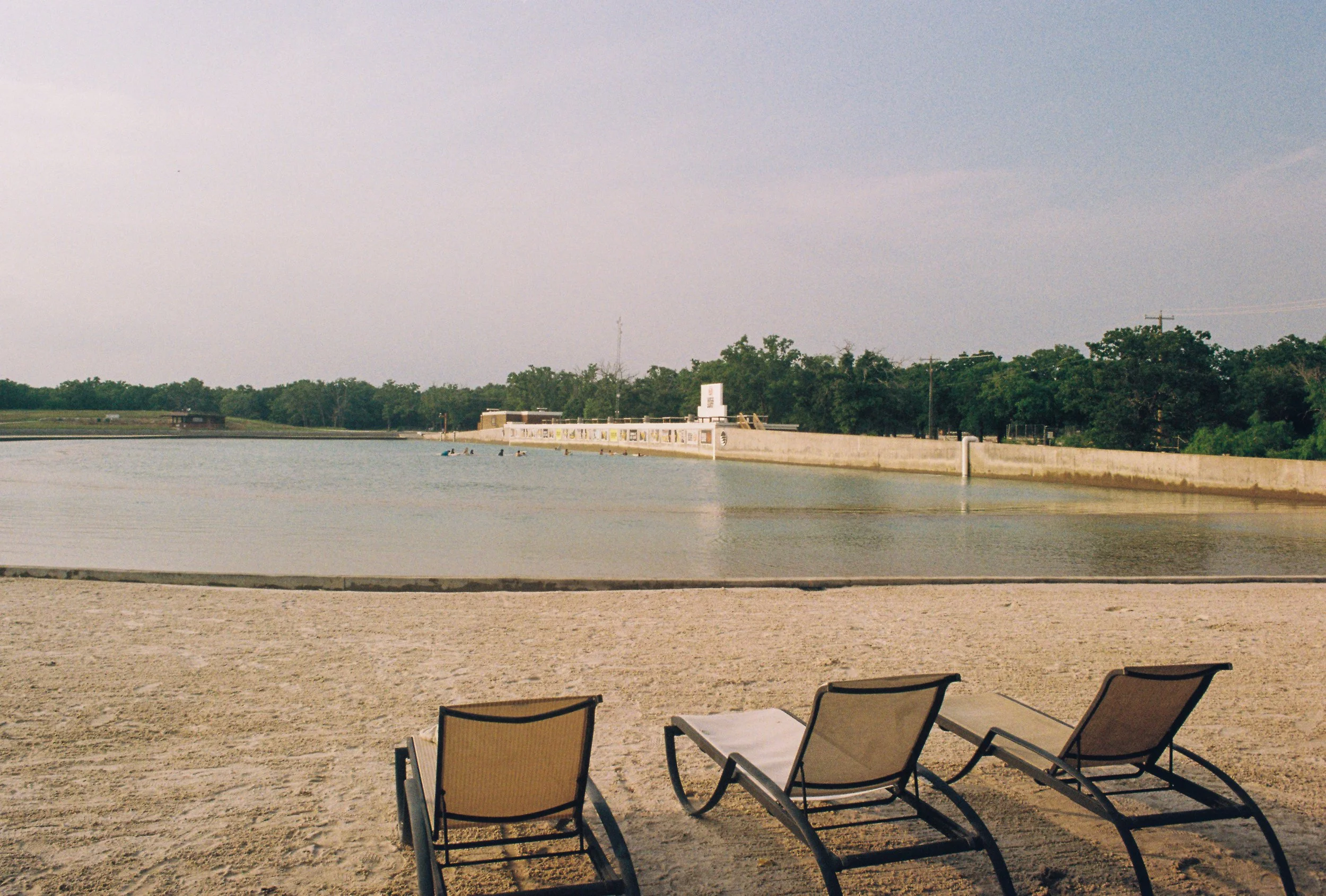 Three empty lounge chairs on a sandy beach facing a calm body of water with a concrete wall and trees in the background.