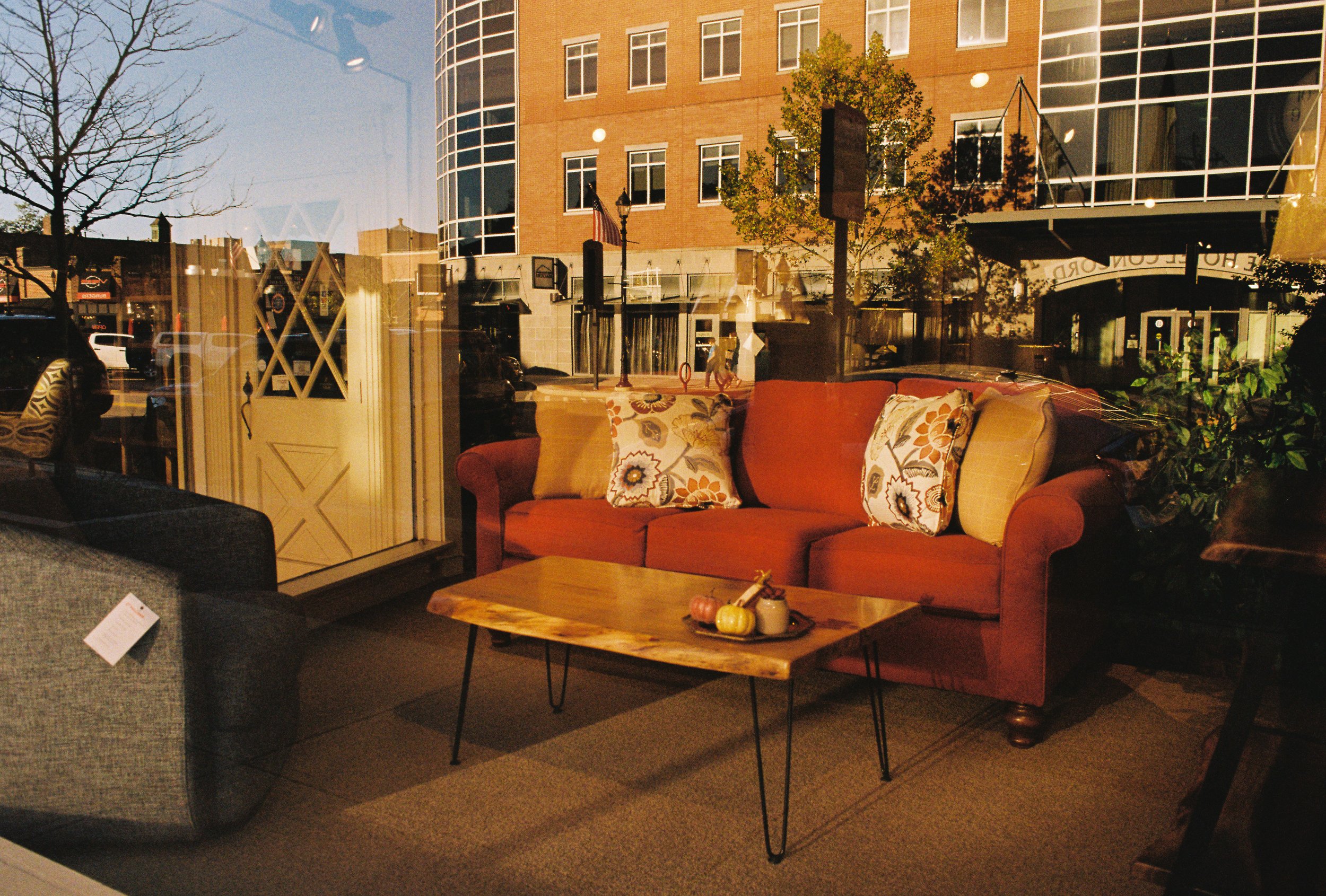 View of a street scene outside a large window showing a modern brick building and trees. Inside, a cozy seating area with a red sofa, patterned pillows, a wooden coffee table with pumpkins and a small jar, and another gray armchair.