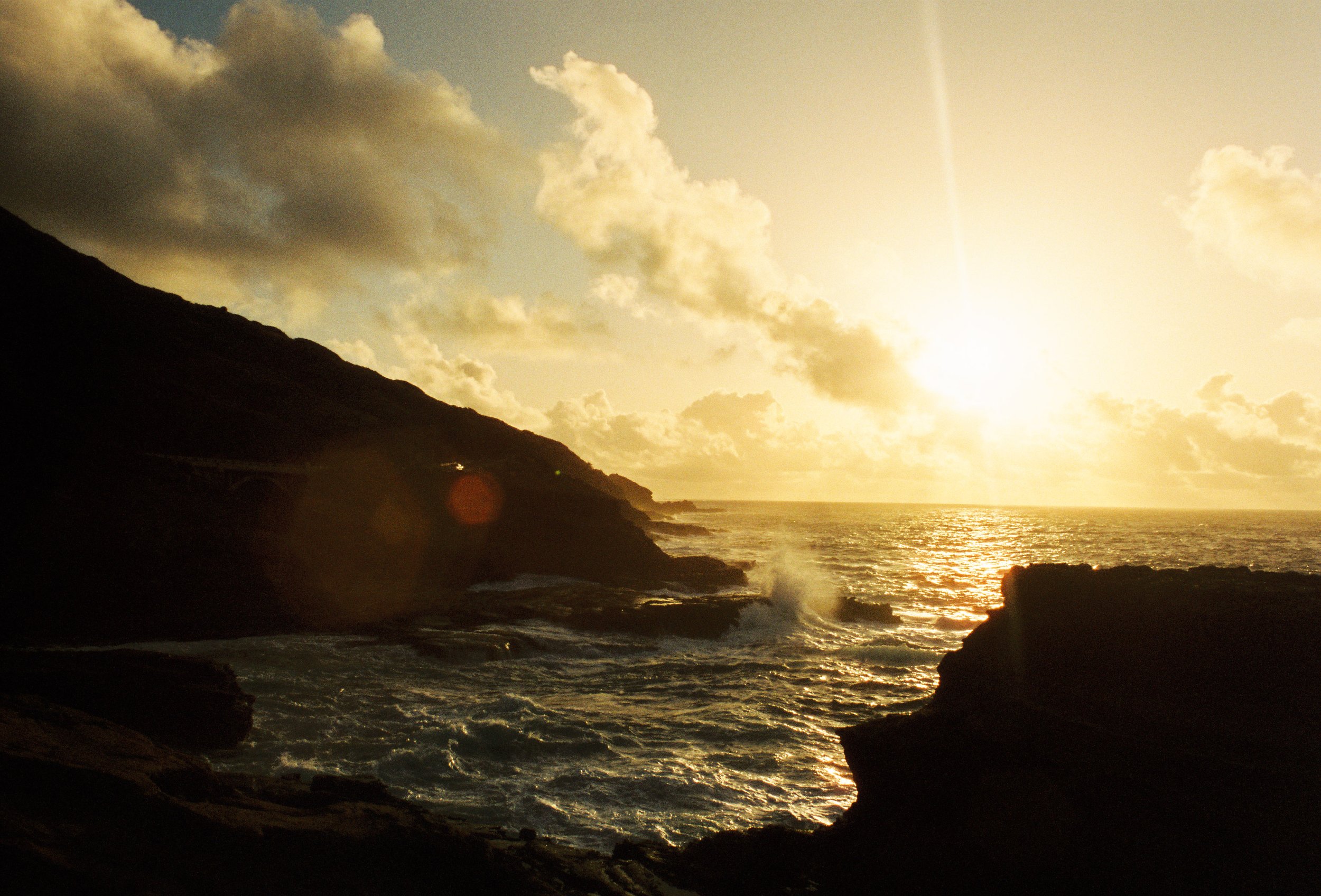 Sunset over the ocean with clouds and silhouetted rocky shoreline.