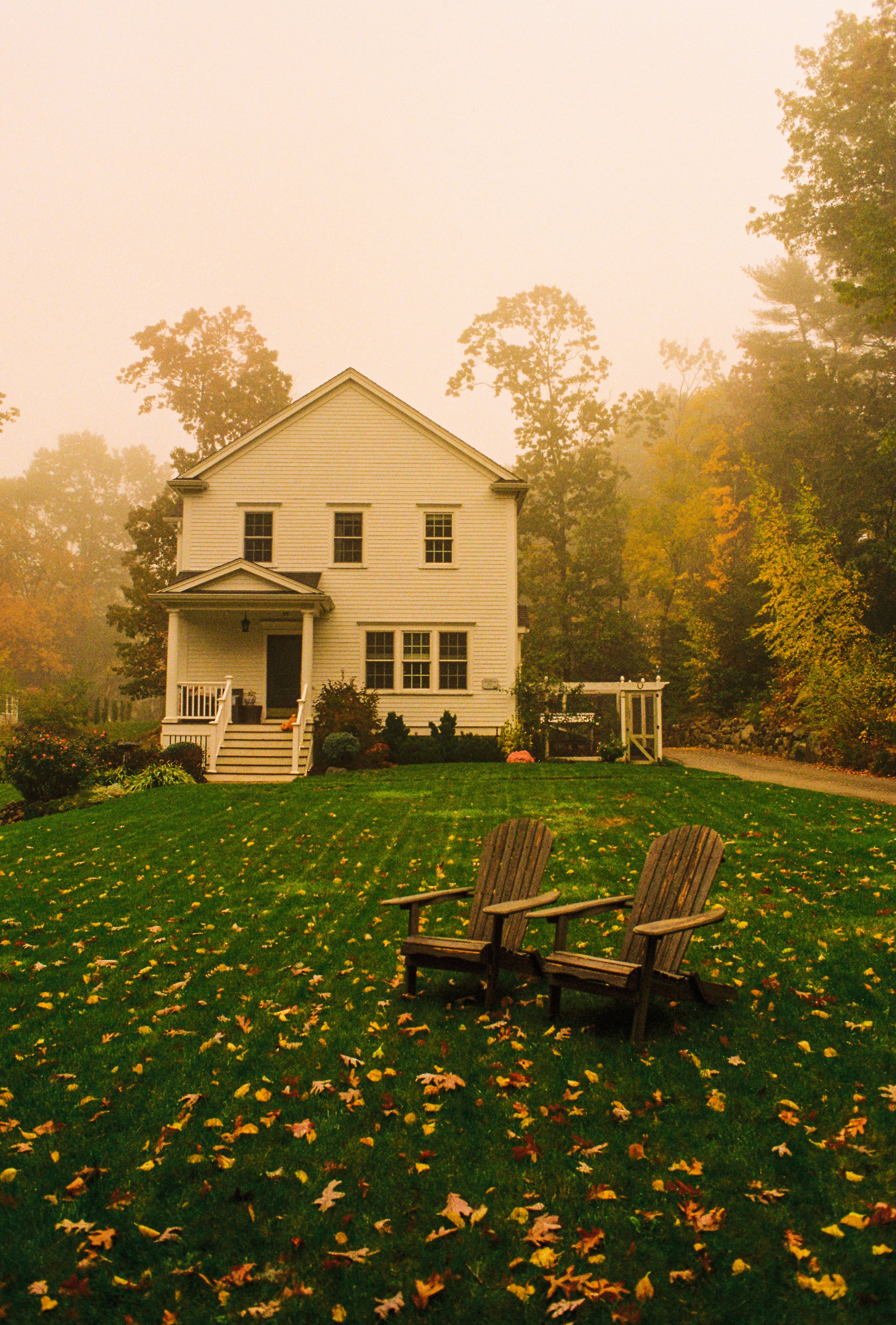 A foggy autumn scene of a white two-story house with a front porch, surrounded by trees and fallen leaves, with two wooden rocking chairs on the lawn.