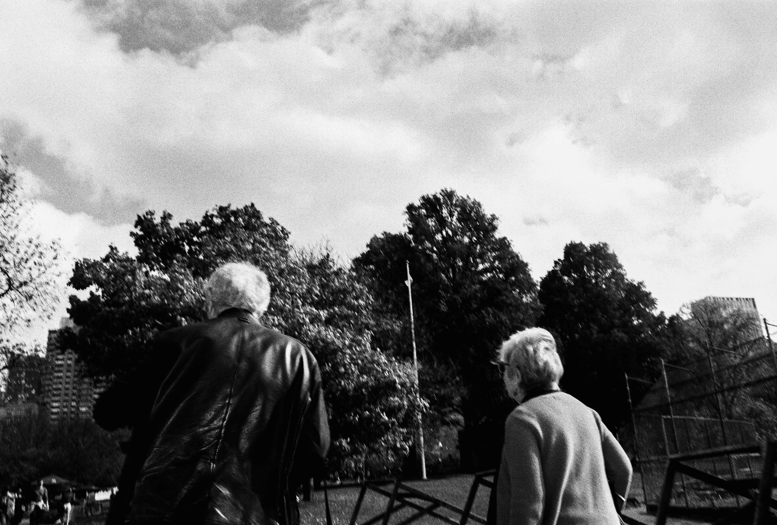 Black-and-white photo of two elderly people, a man and a woman, walking outdoors in a park with trees and buildings in the background.