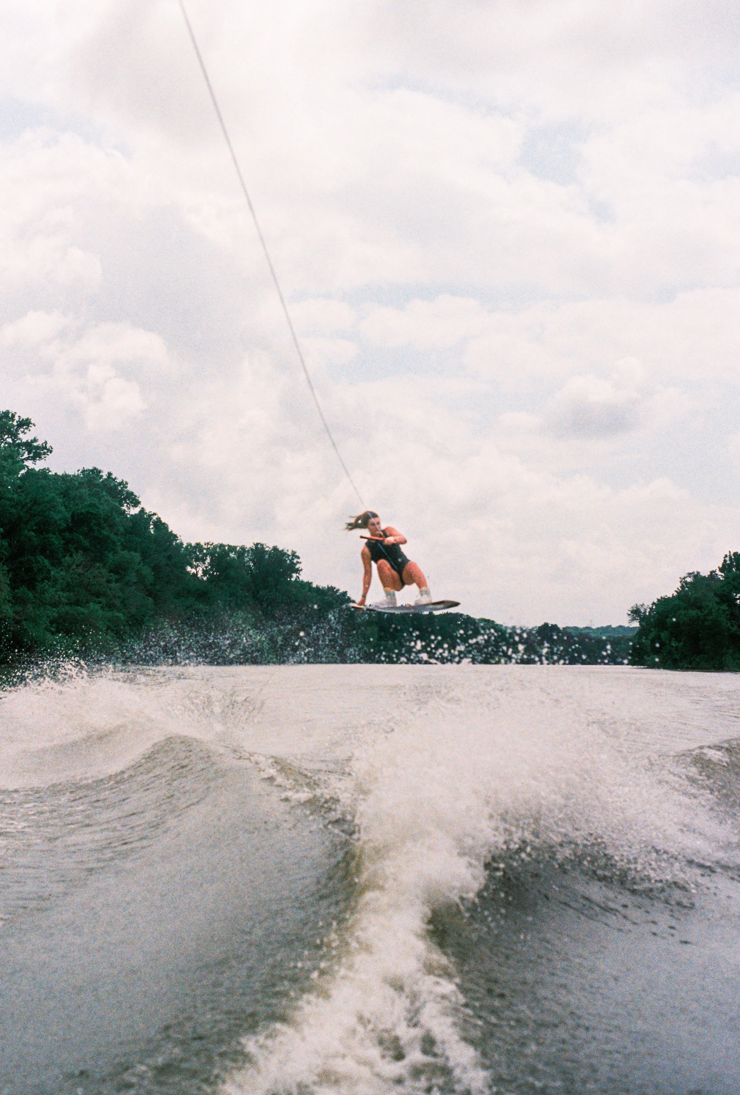 A woman wakeboarding on a lake, holding onto a tow rope, with trees and a cloudy sky in the background.