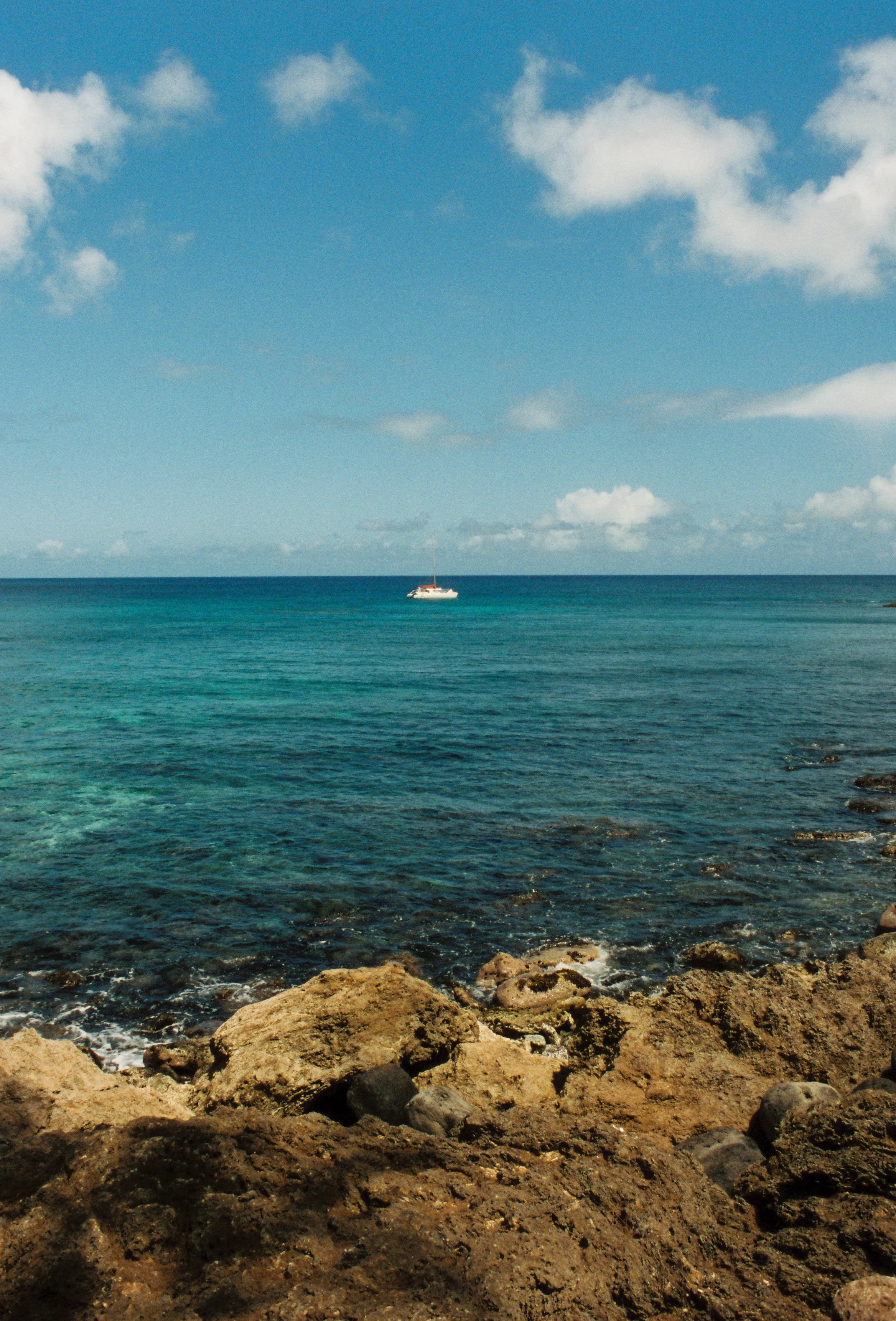 A rocky shoreline with clear blue water and a sailboat in the distance under a partly cloudy sky.