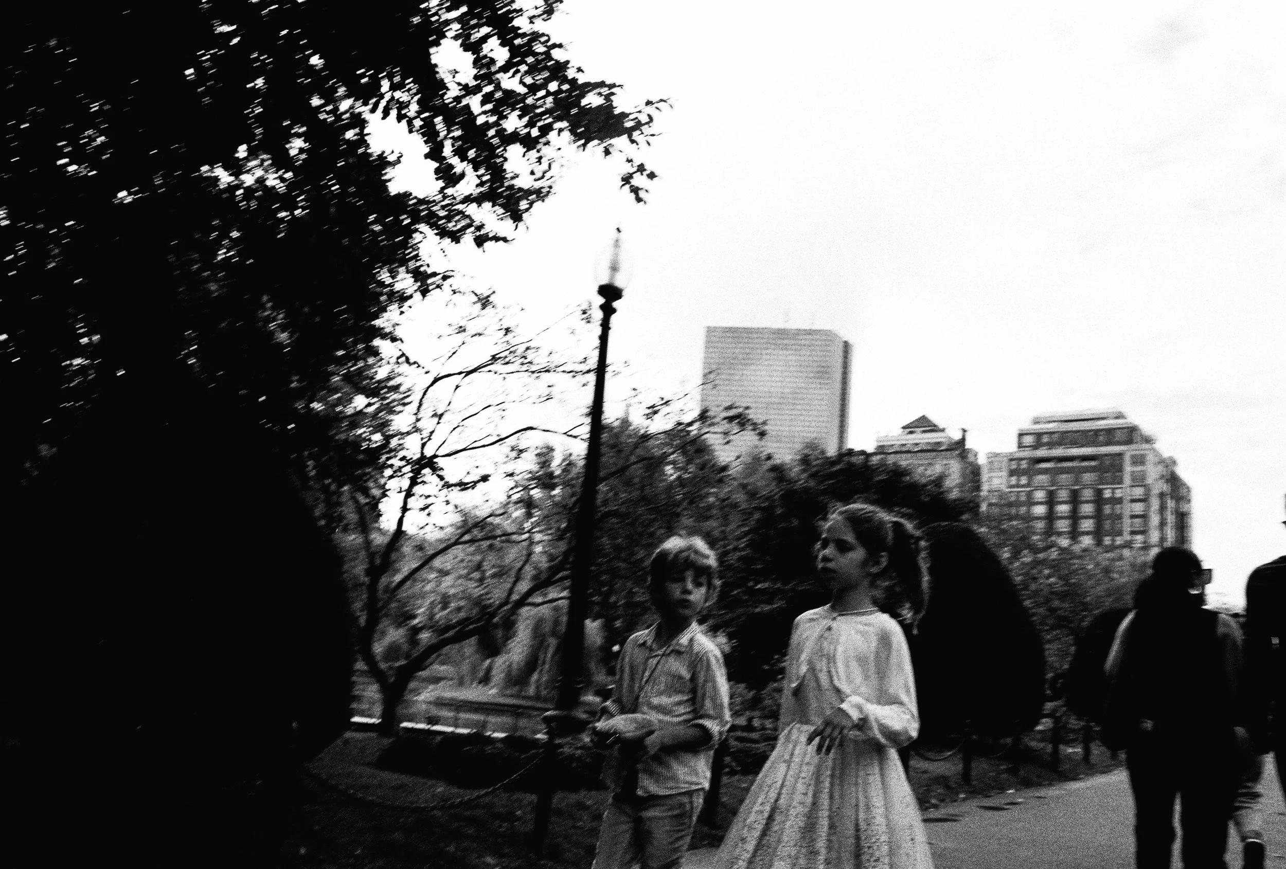 Two children, a boy and a girl, walking in a city park, with buildings and trees in the background, in black and white.