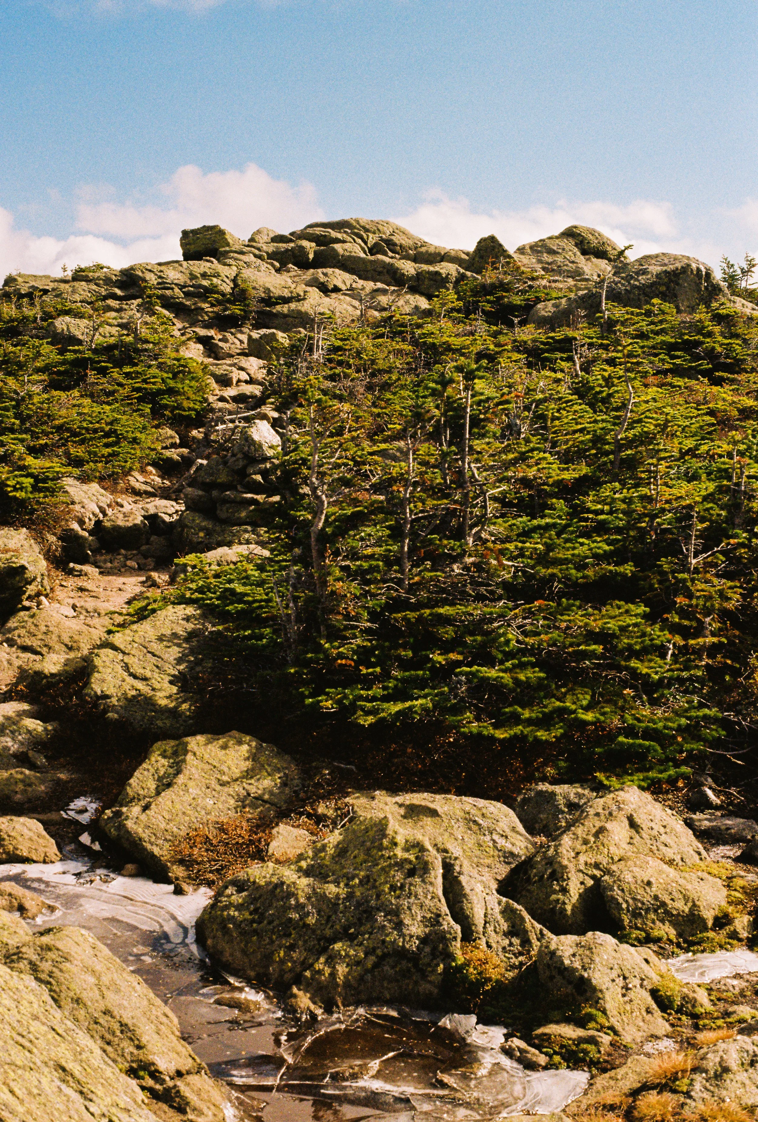 A rugged mountain scene with large rocks, green trees, and a small frozen stream at the bottom, under a bright blue sky with some clouds.