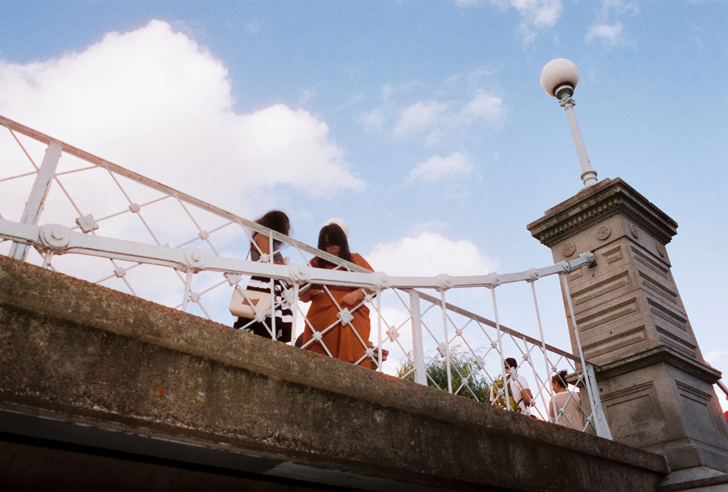 People walking on a bridge with white railing, stone pillars, and a spherical lamp on a cloudy day.