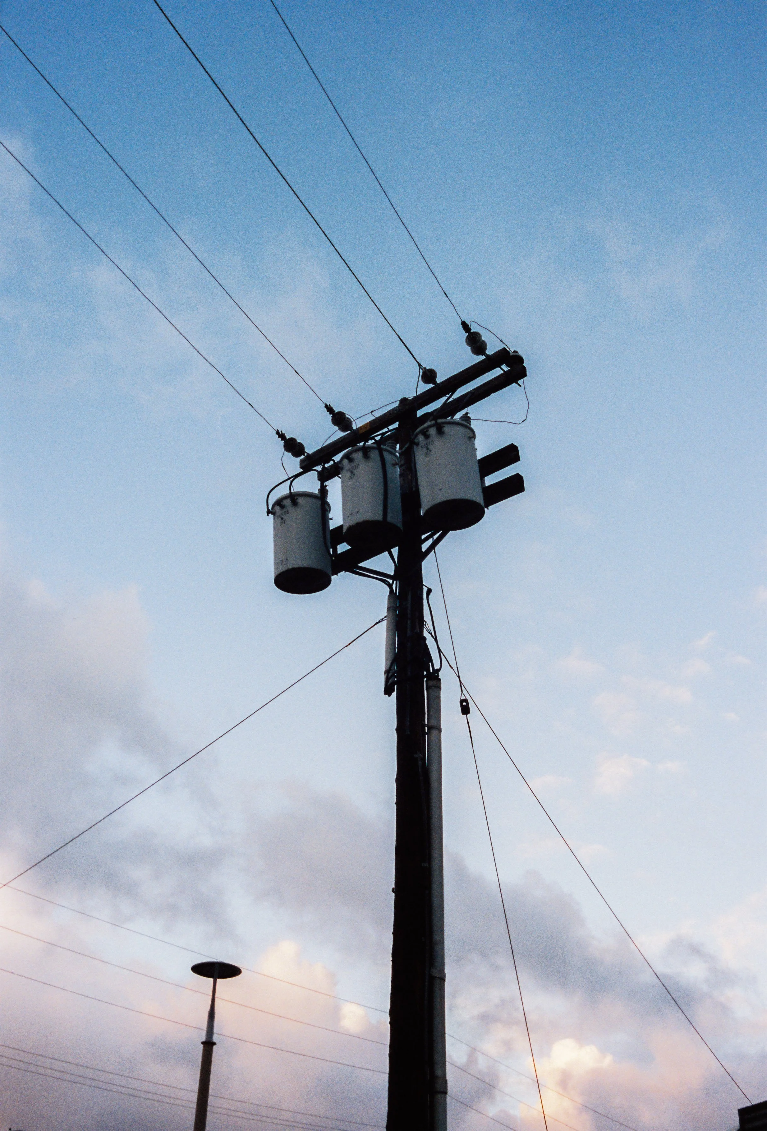 A utility pole with transformers and power lines against a partly cloudy sky at dusk.
