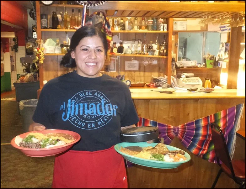 A woman smiling and holding two plates of Mexican food inside a restaurant with a bar in the background.