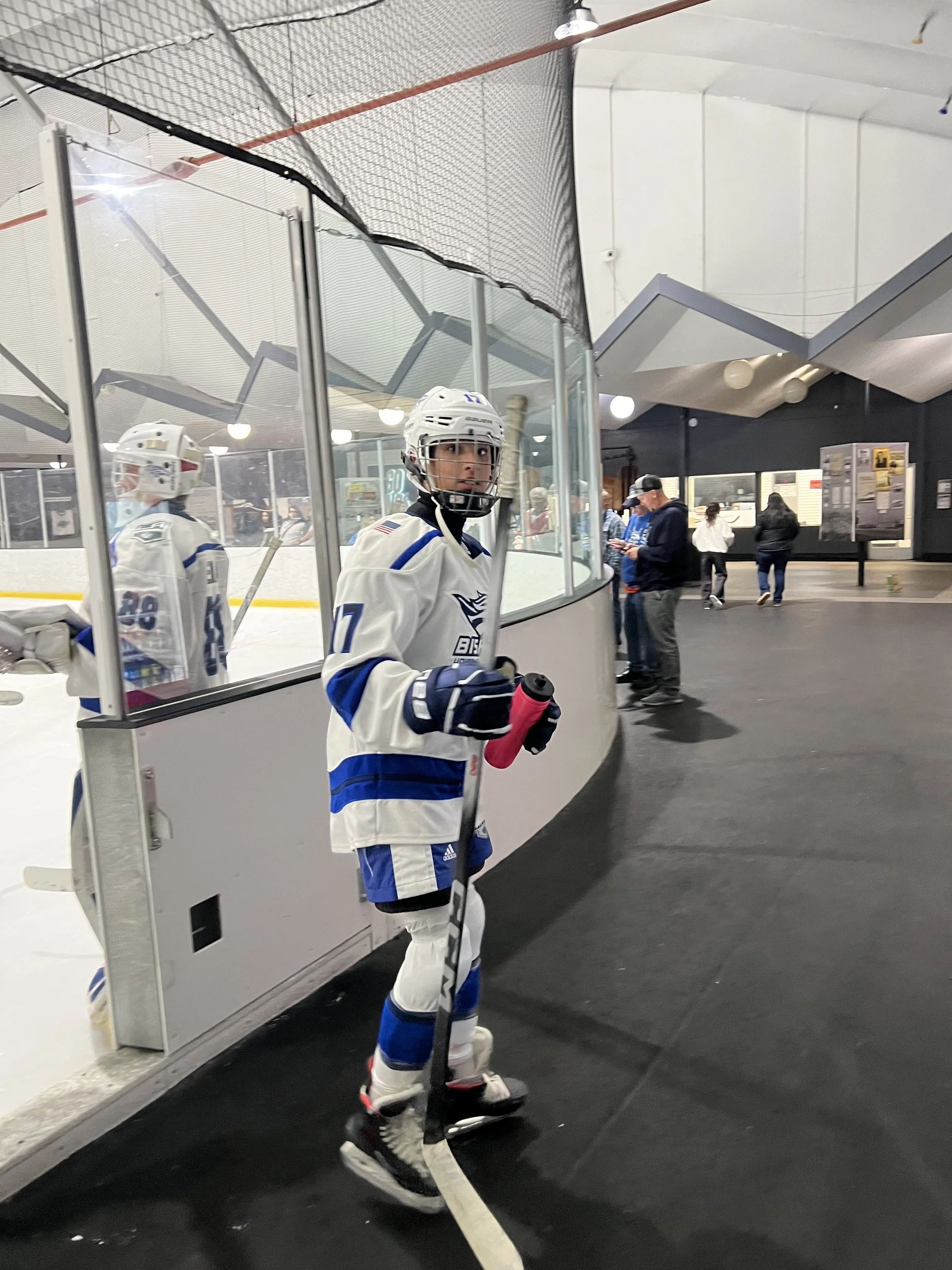 Hockey player in white and blue uniform on the ice rink, holding a pink hockey stick, standing near the bench.