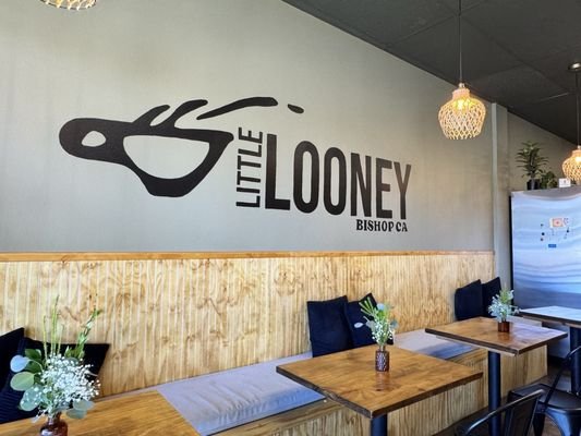 Interior of a restaurant with wooden tables, potted plants, and a large wall decal featuring a guitar and the words "Little Looney" and "Bishop CA".