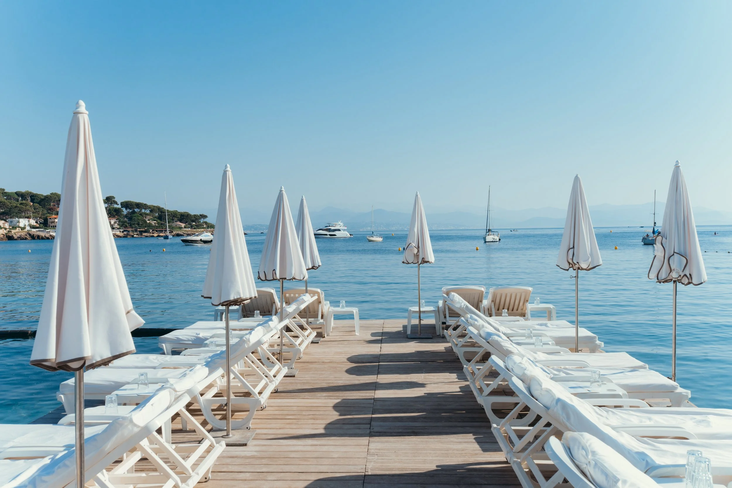 Une Jetée en bois avec des transats blancs et des parasols blancs au bord de la mer, avec plusieurs bateaux à voile et yachts en arrière-plan sous un ciel clair.
