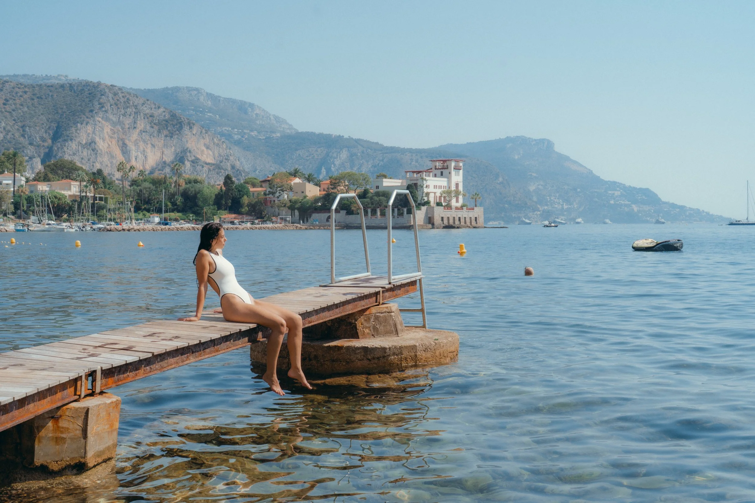 Femme en maillot de bain blanc assise sur un pont en bois au bord de l'eau, avec des montagnes et des bâtiments en arrière-plan, par une journée ensoleillée.