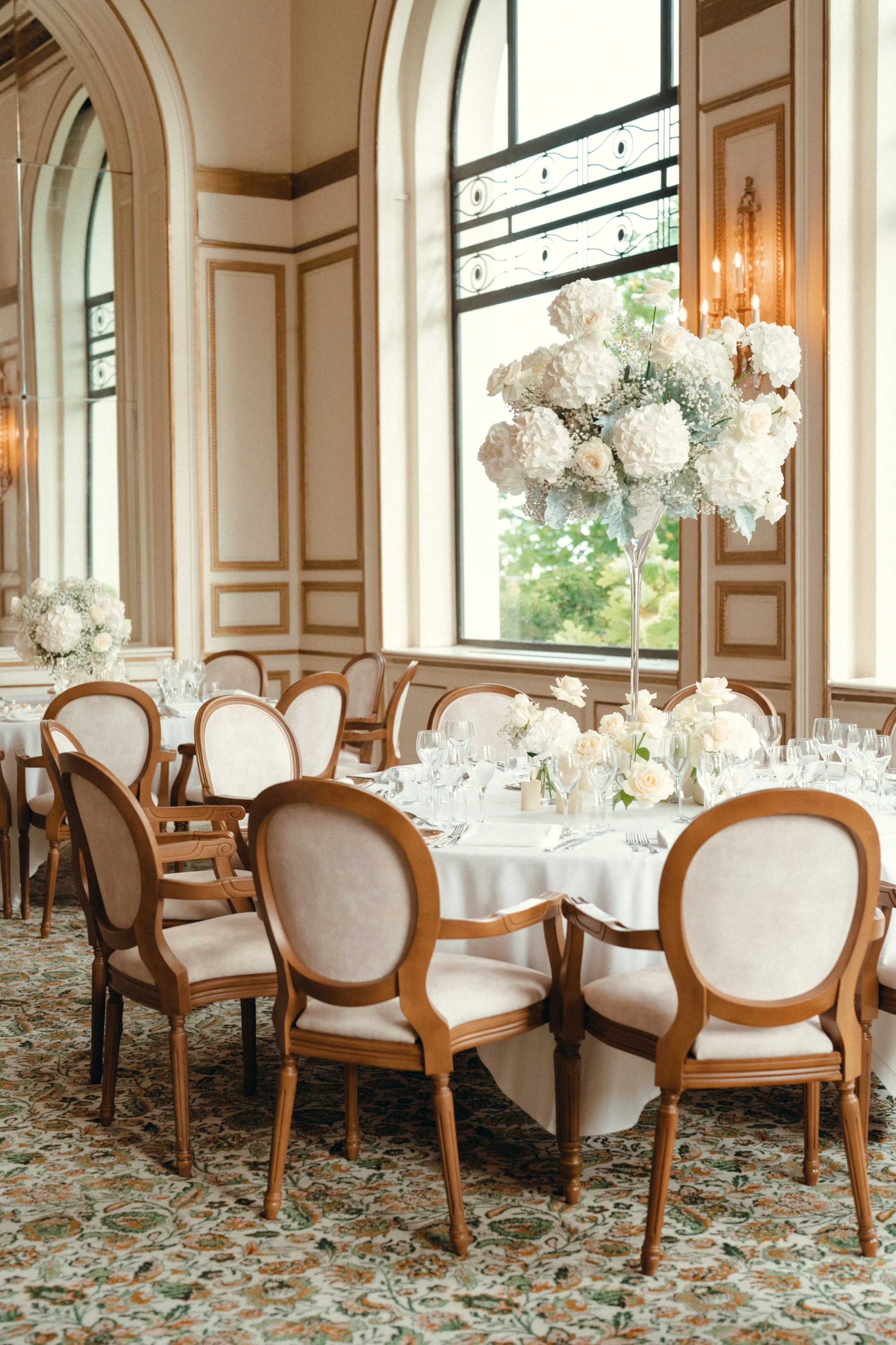 Salle de réception élégante avec une grande table ronde décorée de fleurs blanches, entourée de chaises en bois clair avec des coussins beige, grandes fenêtres laissant entrer la lumière naturelle, et murs ornés en style classique avec moulures et mi