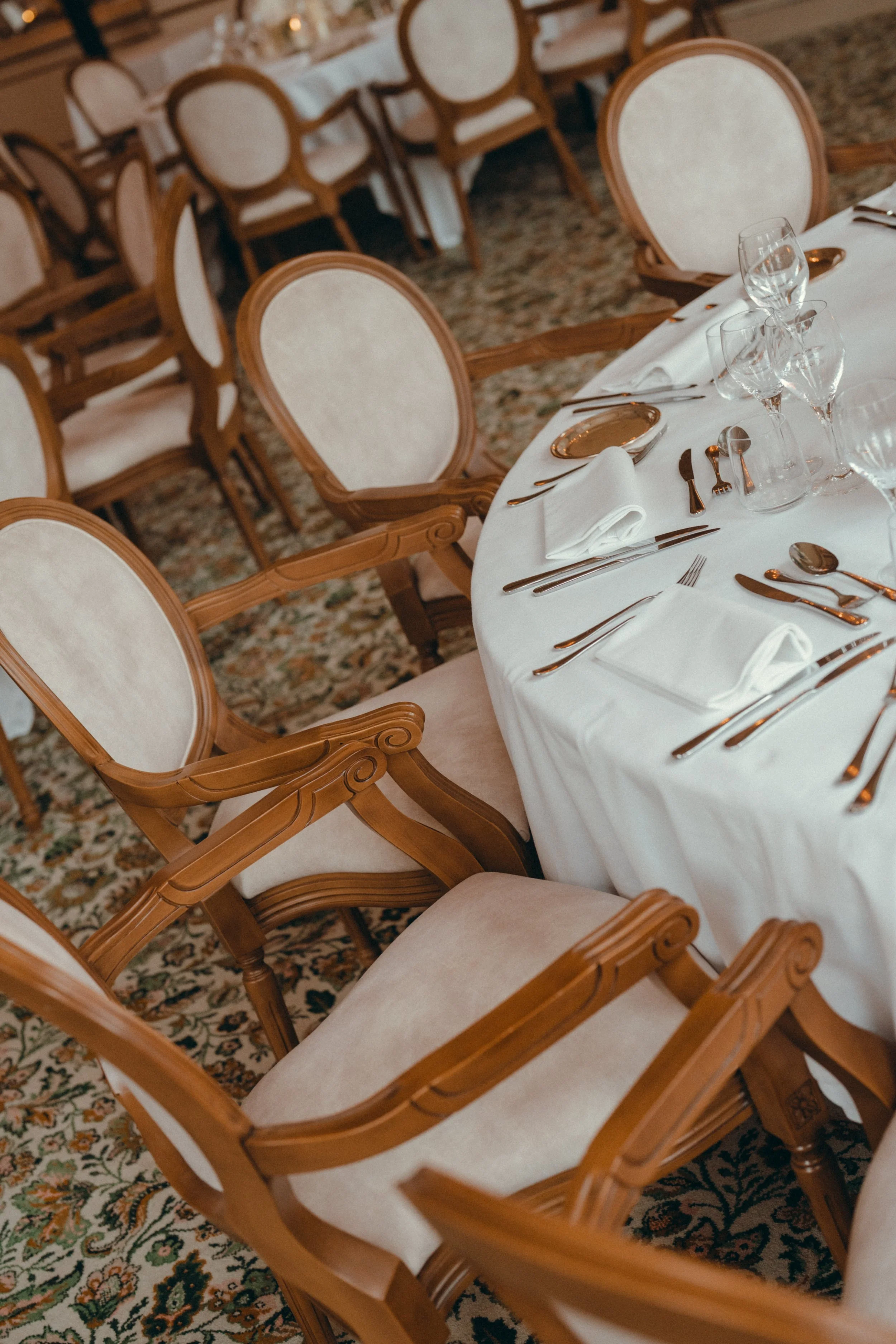 Salle à manger élégante avec une table dressée avec des assiettes, des verres à vin, des couverts en argent et des serviettes pliées. Chaises en bois beige avec des cadres sculptés entourent la table. Le sol est recouvert d'un tapis à motif floral.
