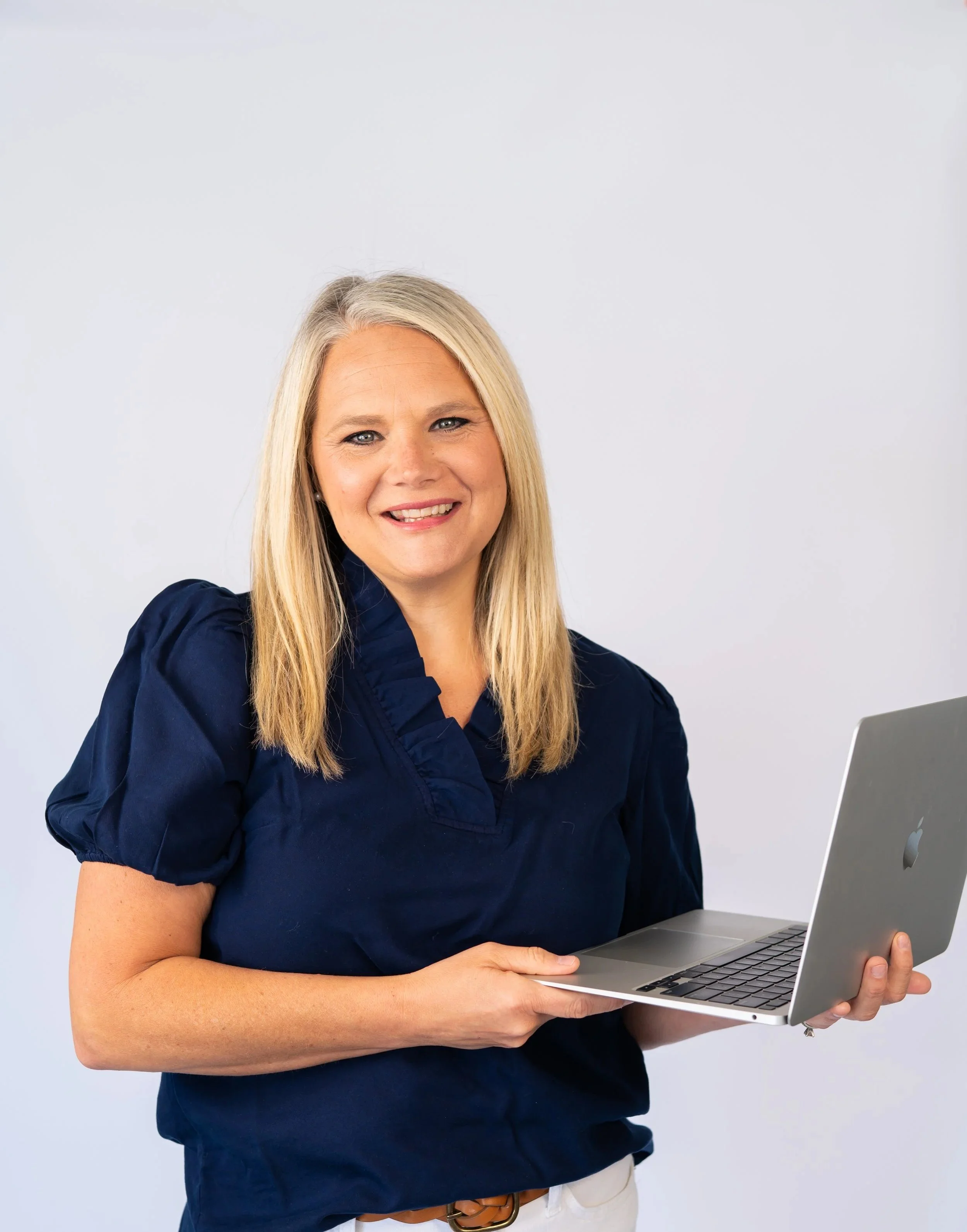 A woman with blonde hair smiling, wearing a navy blue top, holding an open silver Apple MacBook laptop, against a plain light gray background.