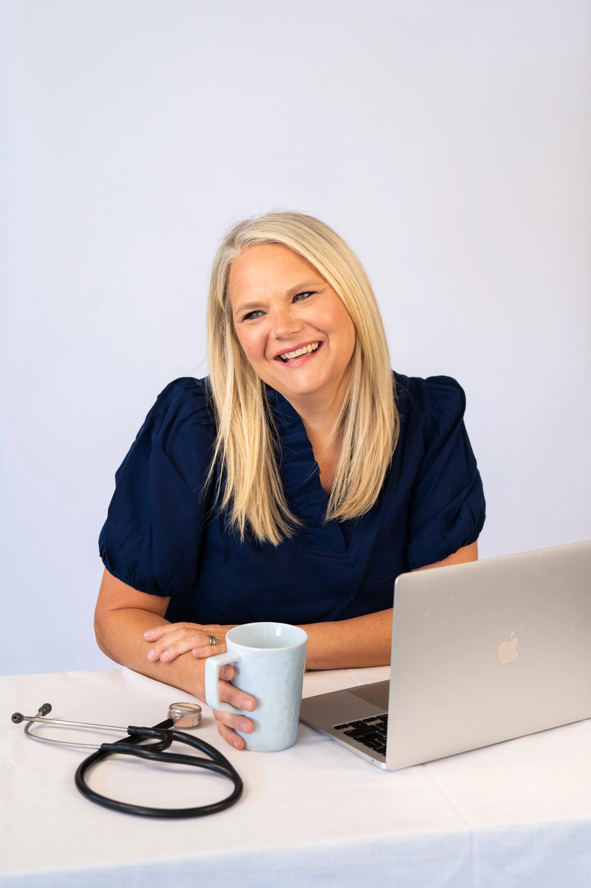 Koral Worsham of Redeeming Roots Functional Wellness sitting at a desk, smiling with a mug in hand, laptop and stethoscope.