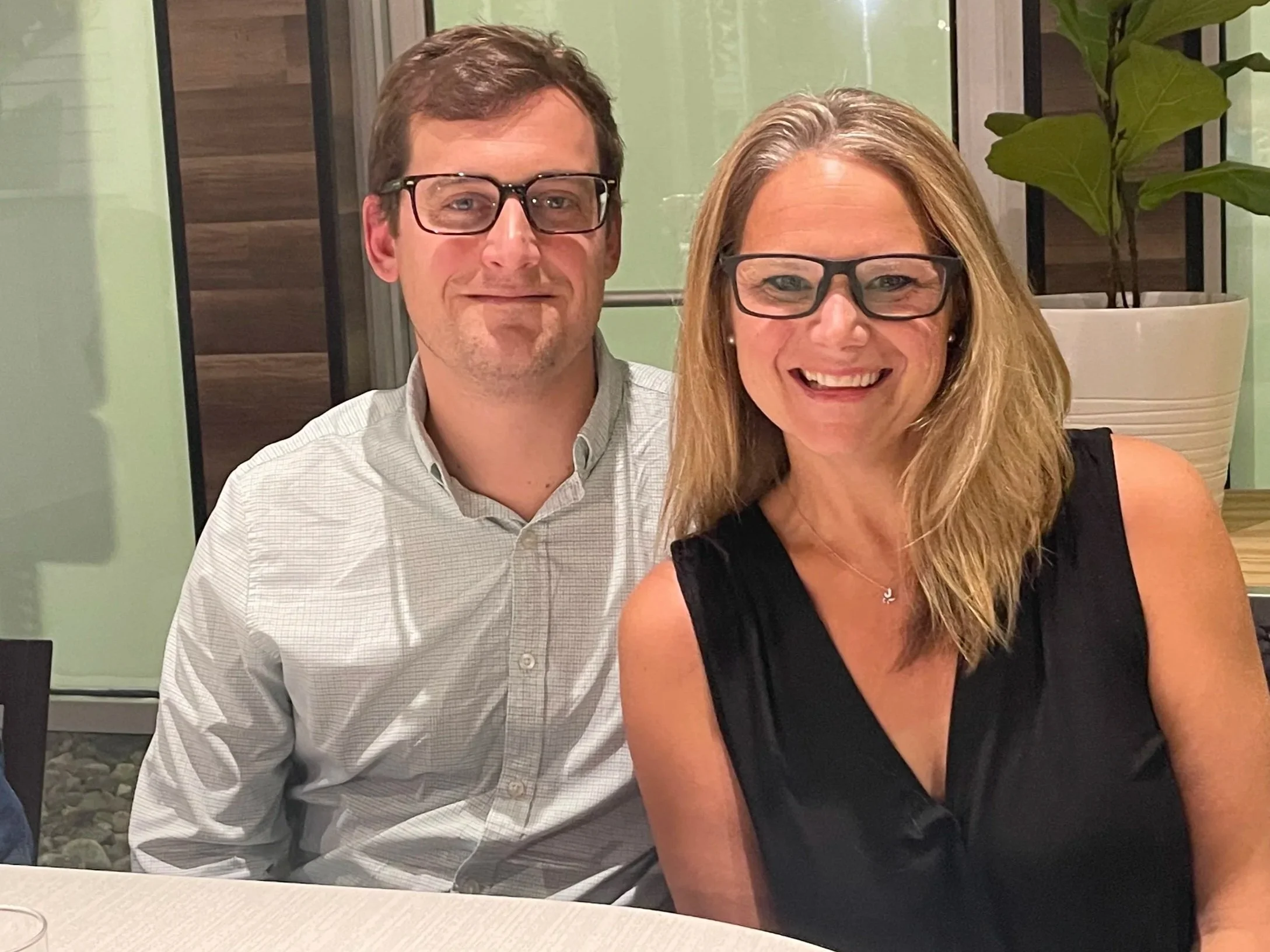 A man and a woman sitting together at a table, smiling for a photo in an indoor setting with a potted plant in the background.