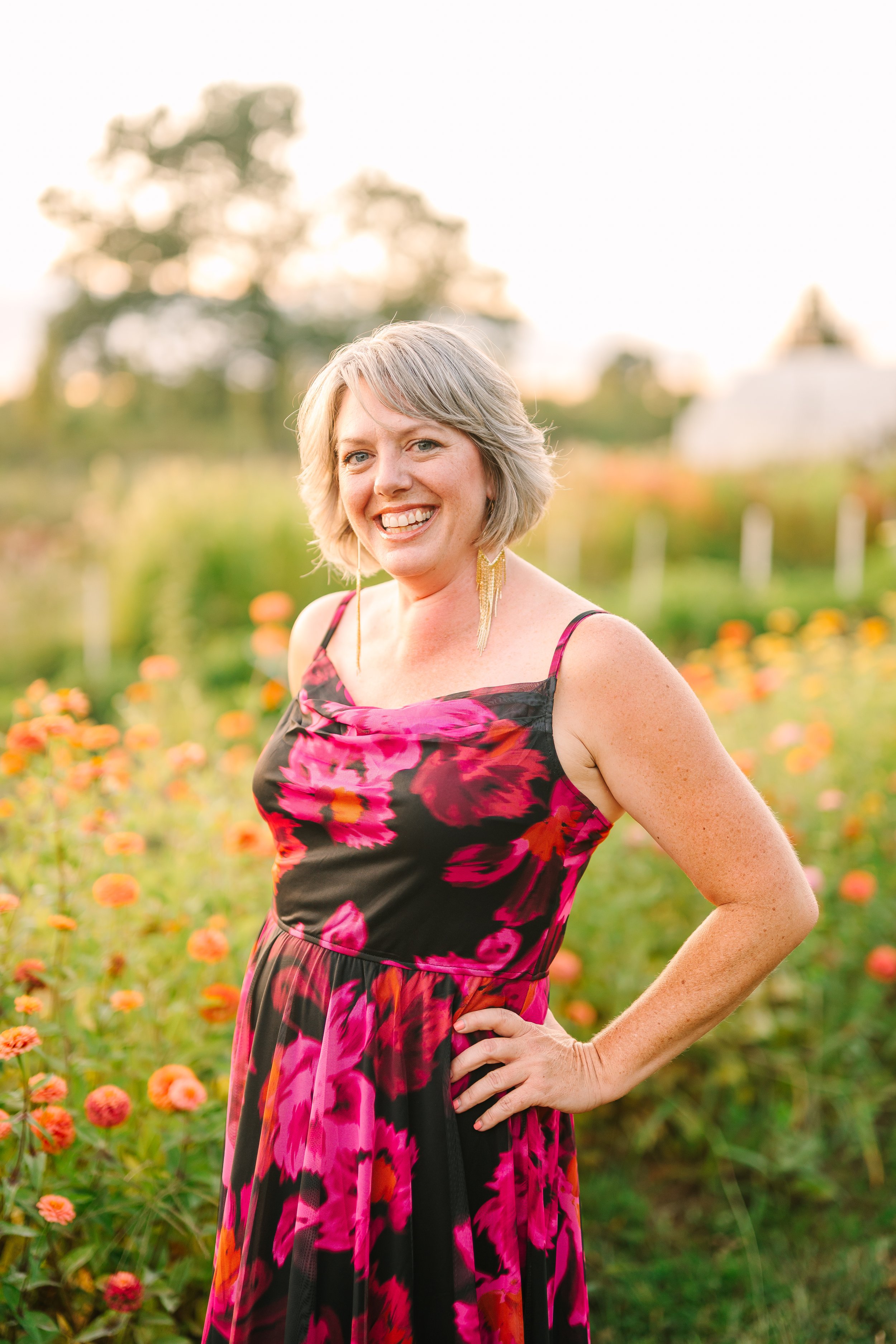 A woman with blond hair wearing a sleeveless maroon dress, smiling outdoors in a forested area.
