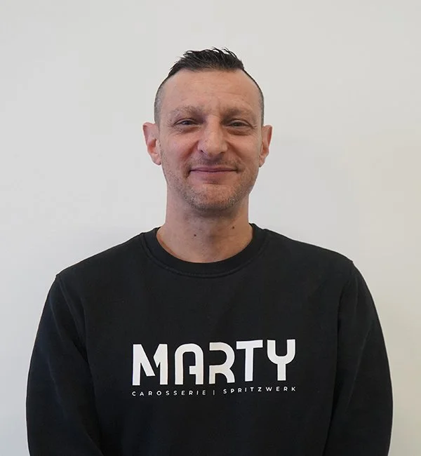 Mann trägt schwarzen Pullover mit weißen Schriftzug 'MARTY Carrosserie Spritzwerk' vor einem weißen Hintergrund.