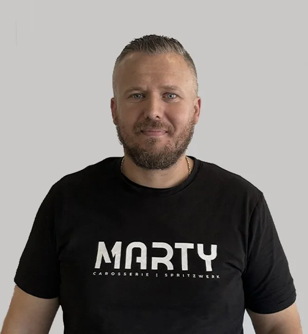Mann mit einem schwarzen T-Shirt mit dem Text "MARTY" und zusätzlichen Worten darunter, vor einem grauen Hintergrund.