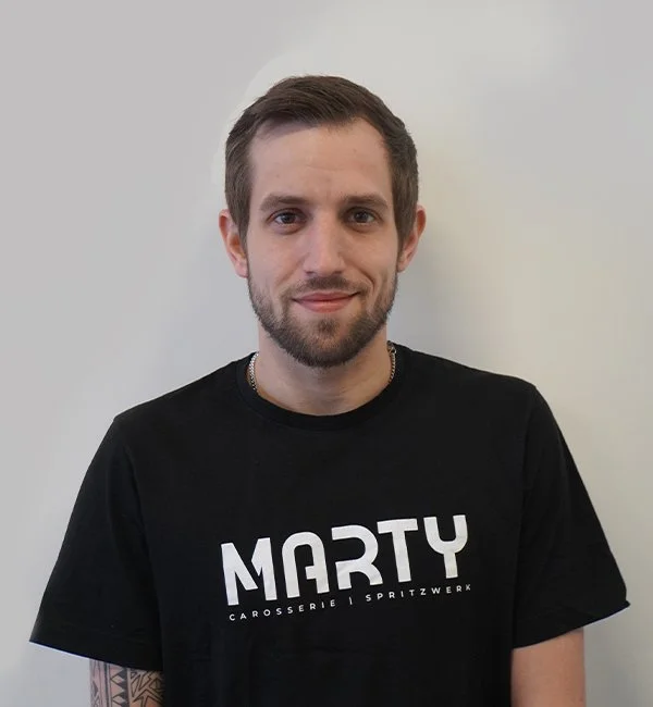 Junger Mann mit dunklem T-Shirt vor weißer Wand, lächelnd.