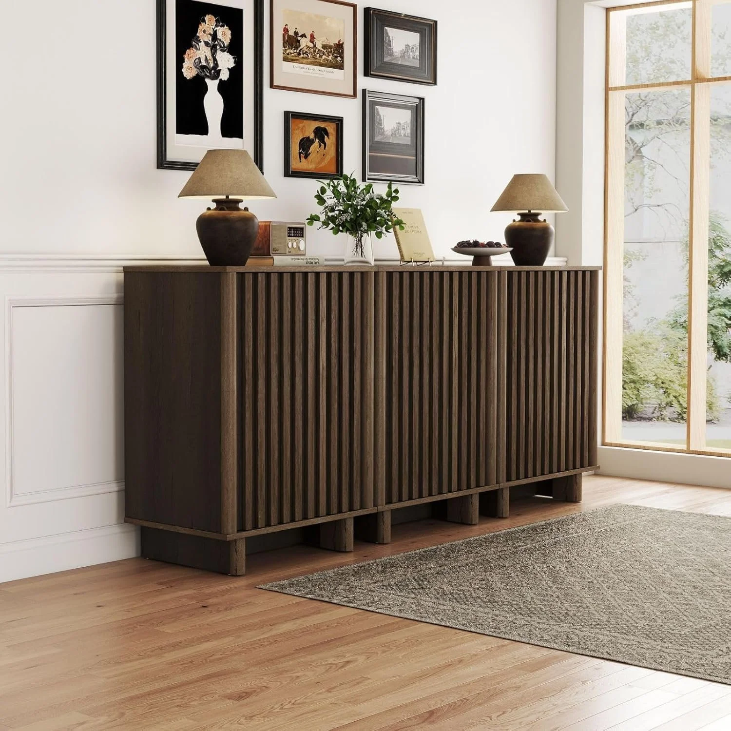 A wooden sideboard with vertical slats on the front, placed against a white wall. On top of it are two table lamps, a small plant, a radio, a book, and a plate of fruit. Above it, a gallery of framed artwork and photos hangs on the wall. To the right, a large window with a view of outdoor greenery.